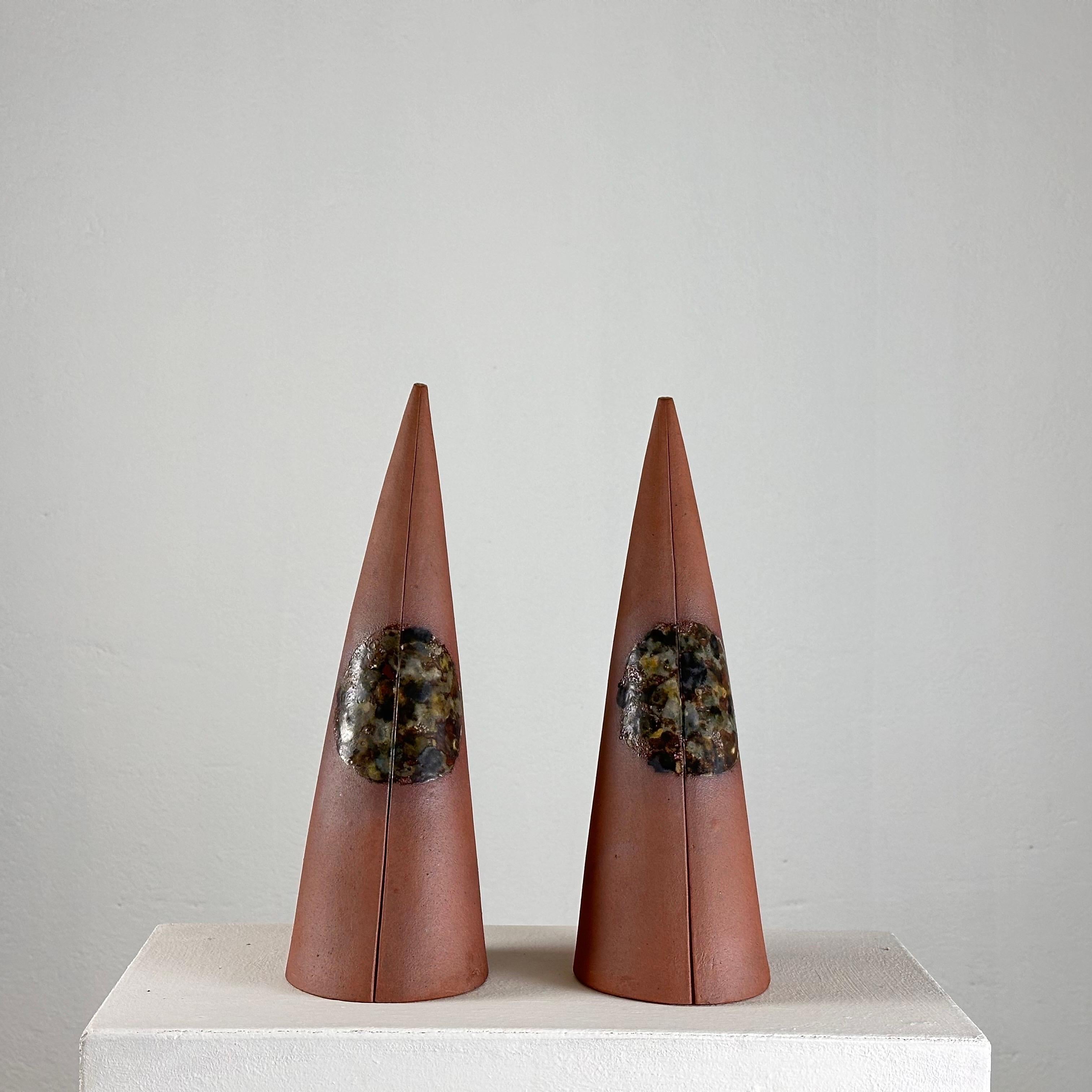 
These cones, signed by the artist, exude a timeless elegance and sophistication. Crafted in a warm terracotta hue and embellished with a spherical motif at the midpoint, reminiscent of the cosmos, they evoke a sense of cosmic harmony and