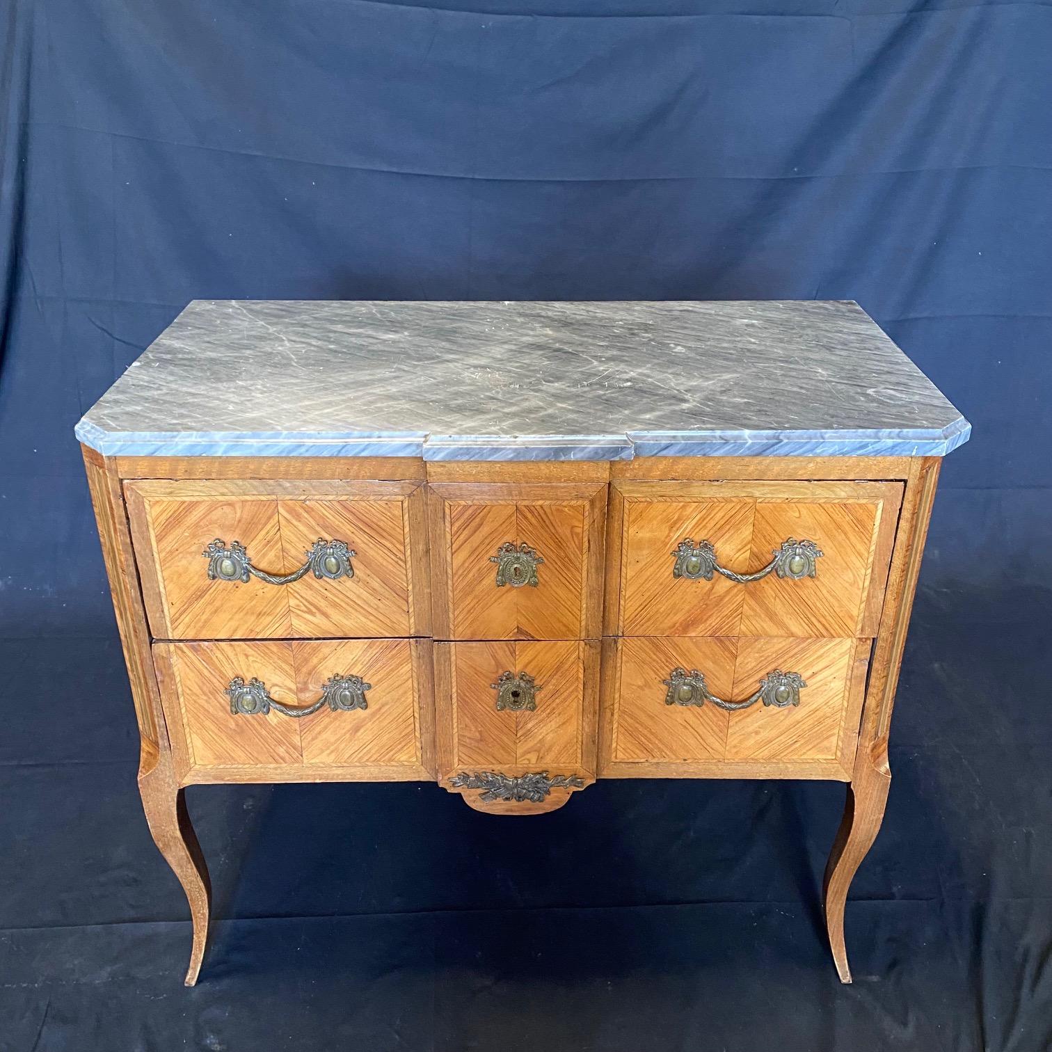 French antique 2 drawer commode having exquisite attention to detail with fine floral marquetry inlays and a central marquetry vignette with musical instruments accented with a bow. The stunning marble top is original and the bronze mounts crisply