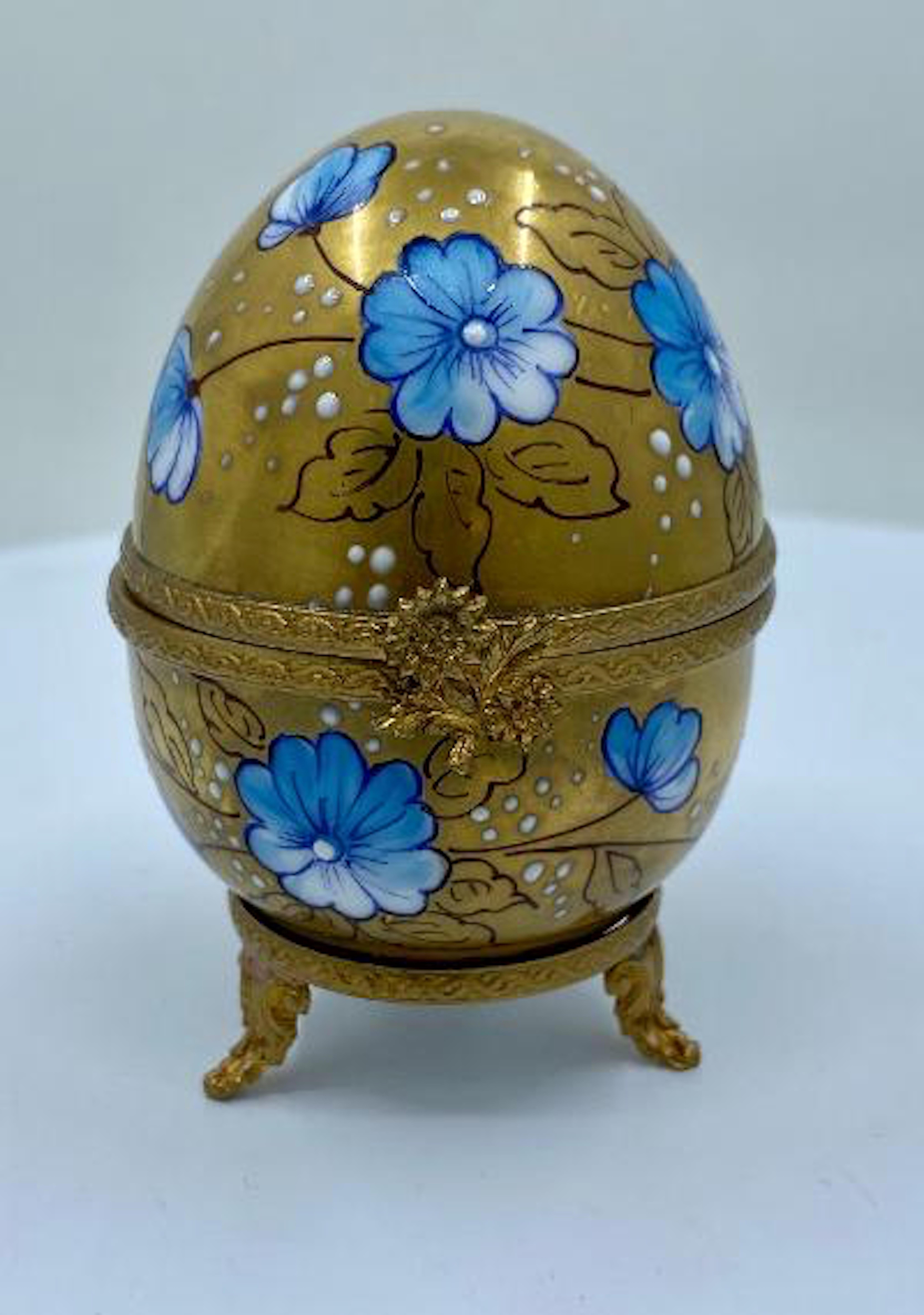 Exquisitely handmade and hand painted miniature Limoges porcelain egg shaped box with a hinged lid is decorated with light blue and white flowers accented with high relief accents over a lavish 24-karat gold background. The porcelain box and lid