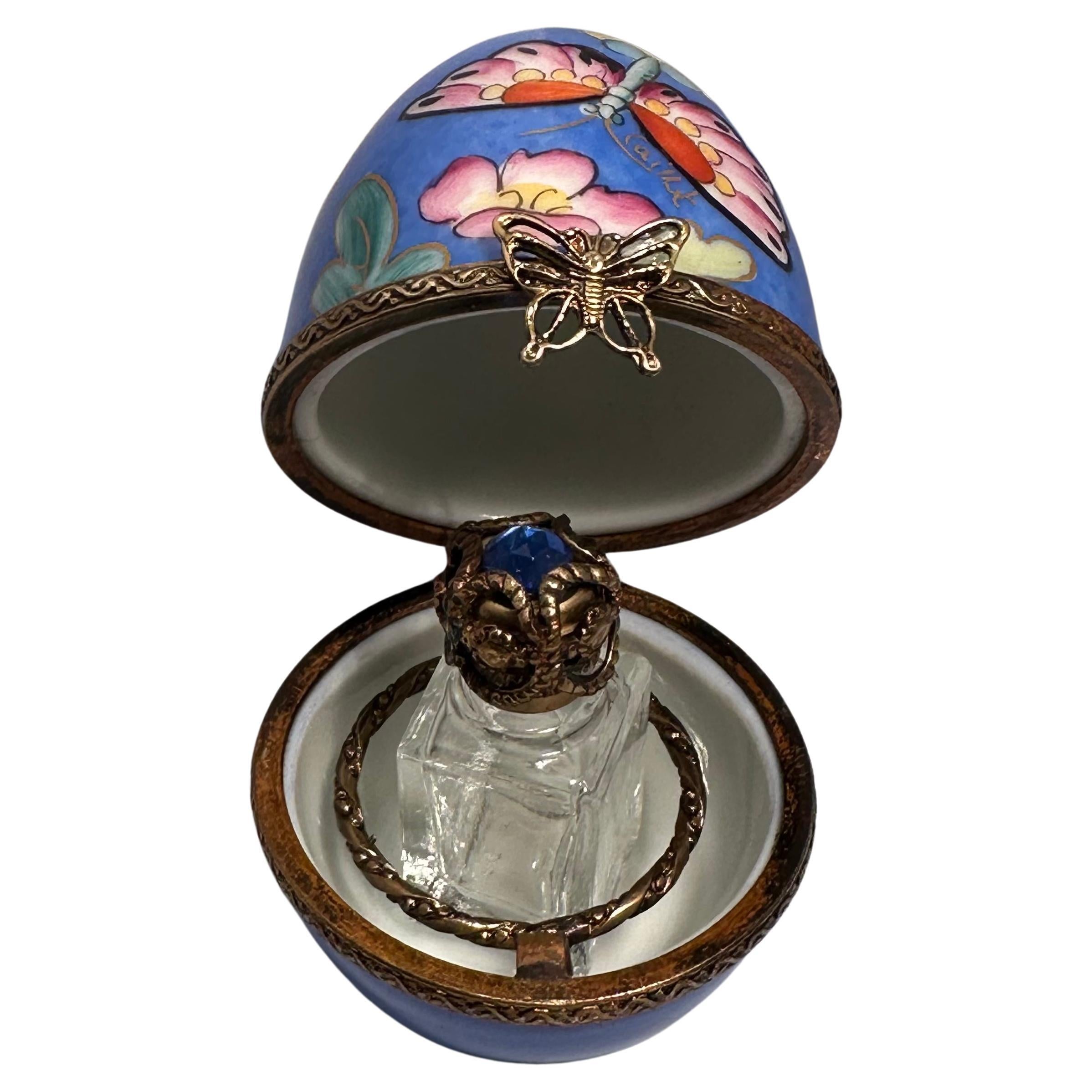 Exquisitely handmade and hand painted miniature Limoges porcelain egg shaped perfume box with a hinged lid is decorated with pretty polychrome flowers and Butterly motif over a soothing blue background. The porcelain box and lid feature rims or