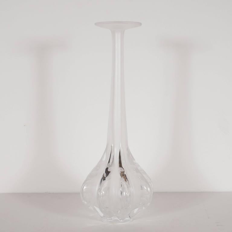 This stunning vase is designed by Marie Claude Lalique. This vase is of clear and frosted glass with a long slender neck with a flaired opening. The base has eight protrusions with wheel enhanced markings and has a polished rim and base. Verso
