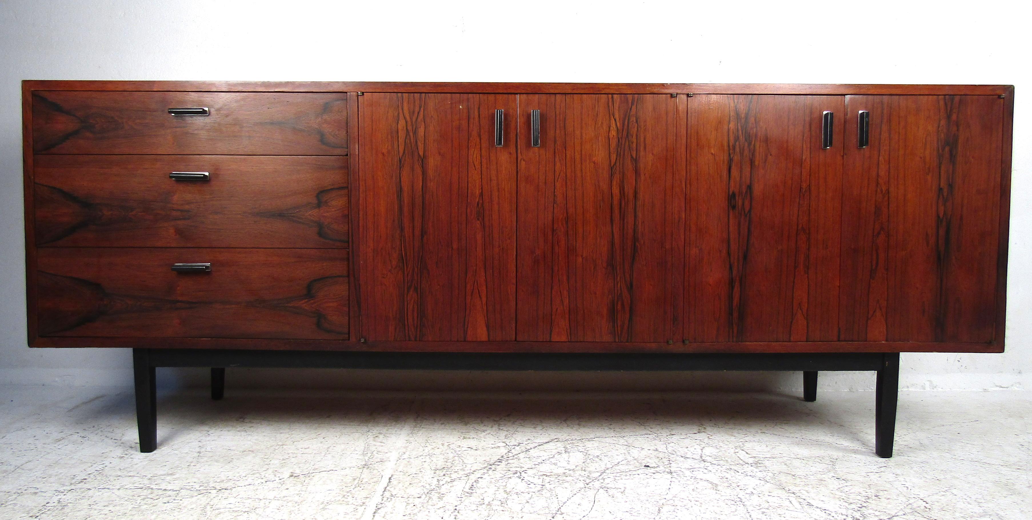 This stunning vintage modern credenza features a beautiful rosewood front with two cabinet doors and three hefty drawers. Ample storage is provided by two large compartments with shelves and drawers. The unusual sculpted door handles and black