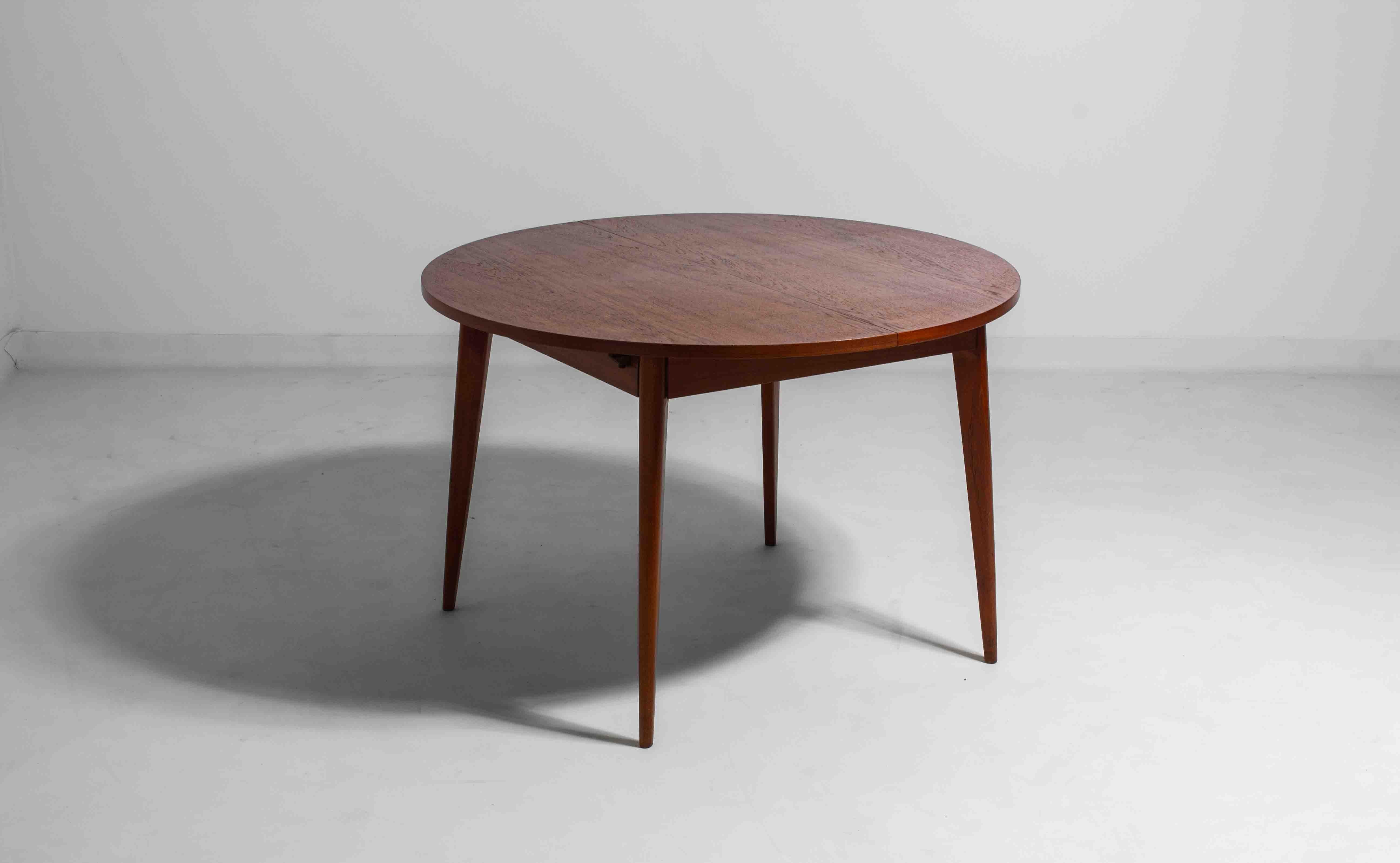 This mid-century modern dining table from the 1960s is made of warm teak and features a simple, round design with tapered legs. It extends with a hidden central leaf, making it easy to go from a compact table to a larger one for gatherings. A