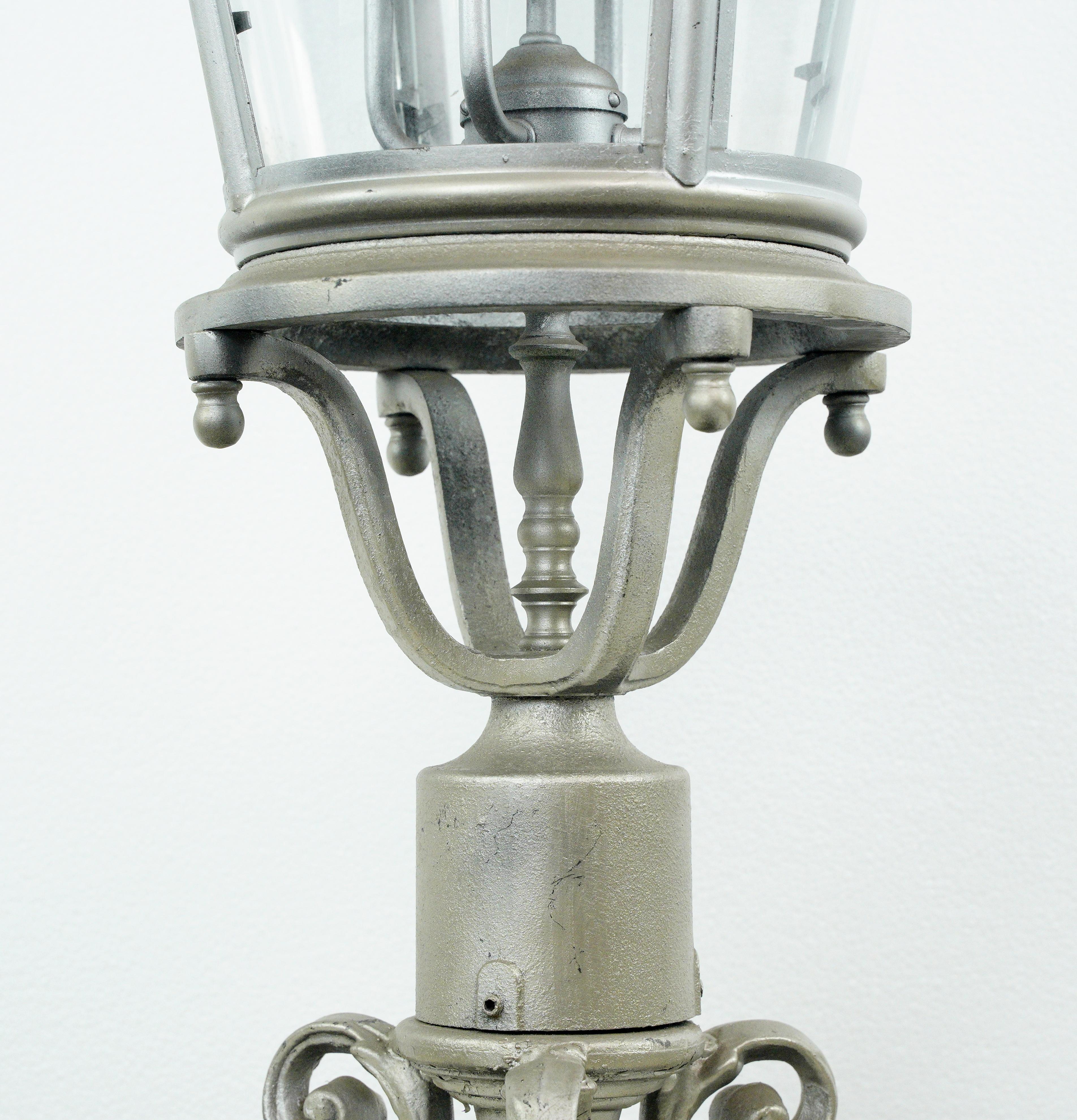 This antique aluminum column or post lantern light is a stunning outdoor fixture with a decorative scroll design at the base, distressed silver paint, and five glass enclosed candelabra arm lights. It combines old world charm with durability,