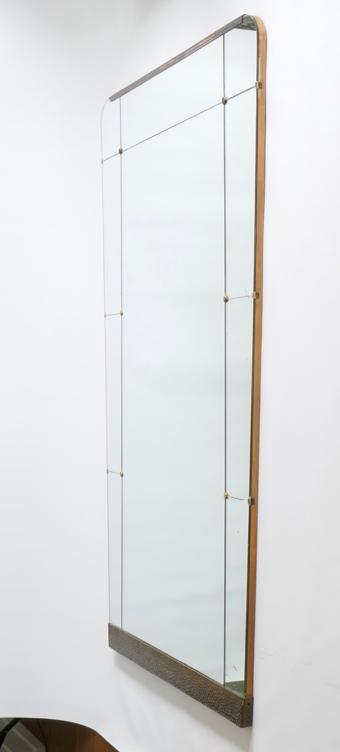 A very large wall mirror of tapering rectangular form, composed of one large central glass within a contour of smaller glass panels, the panels to each top corner gently rounded. Embellished with a hammered brass element to the base and a polished