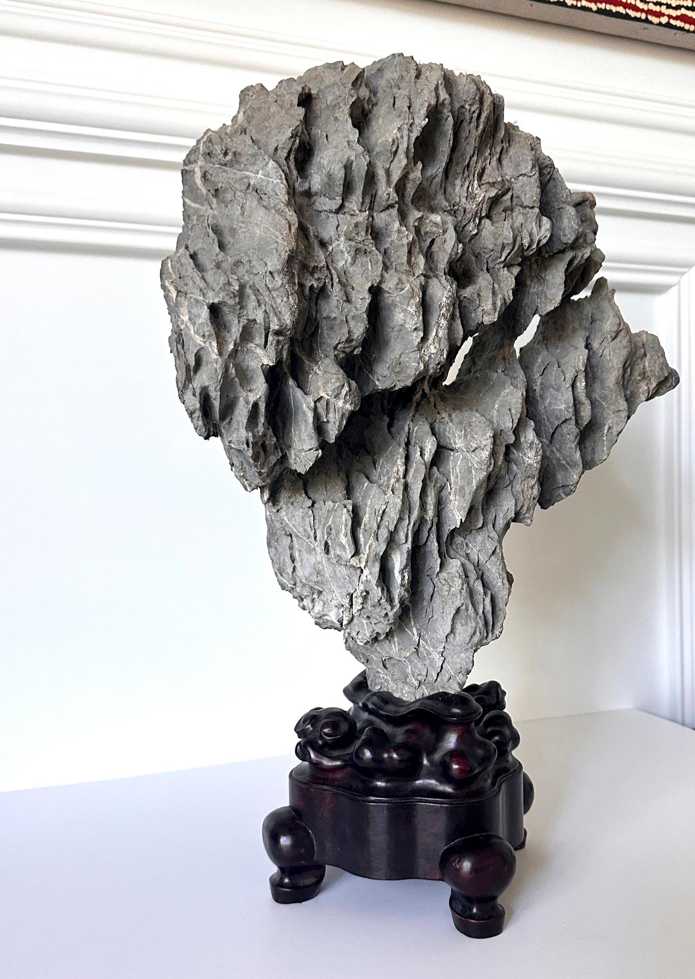 L'objet proposé est un rocher vertical de lettré chinois posé sur un présentoir en bois sculpté. Ce spécimen est un magnifique exemple de pierre Ying (ou Yingde), l'un des quatre célèbres types de pierre connus des lettrés chinois, moins courant que