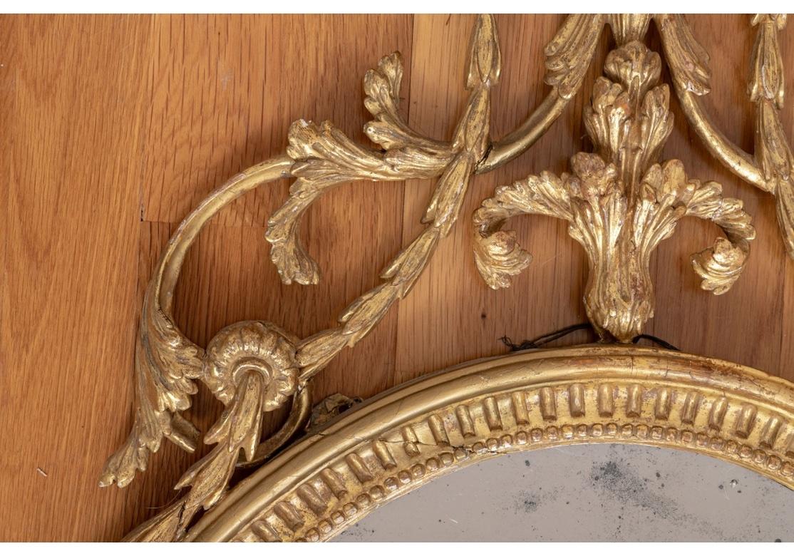 An important Adam influenced design in the tall carved and gilt oval mirror. The tall top element with a pine cone and leafy crest atop a footed urn with bell flower swags. A center fluted horn with scrolling leaf and bell flower vines are attached