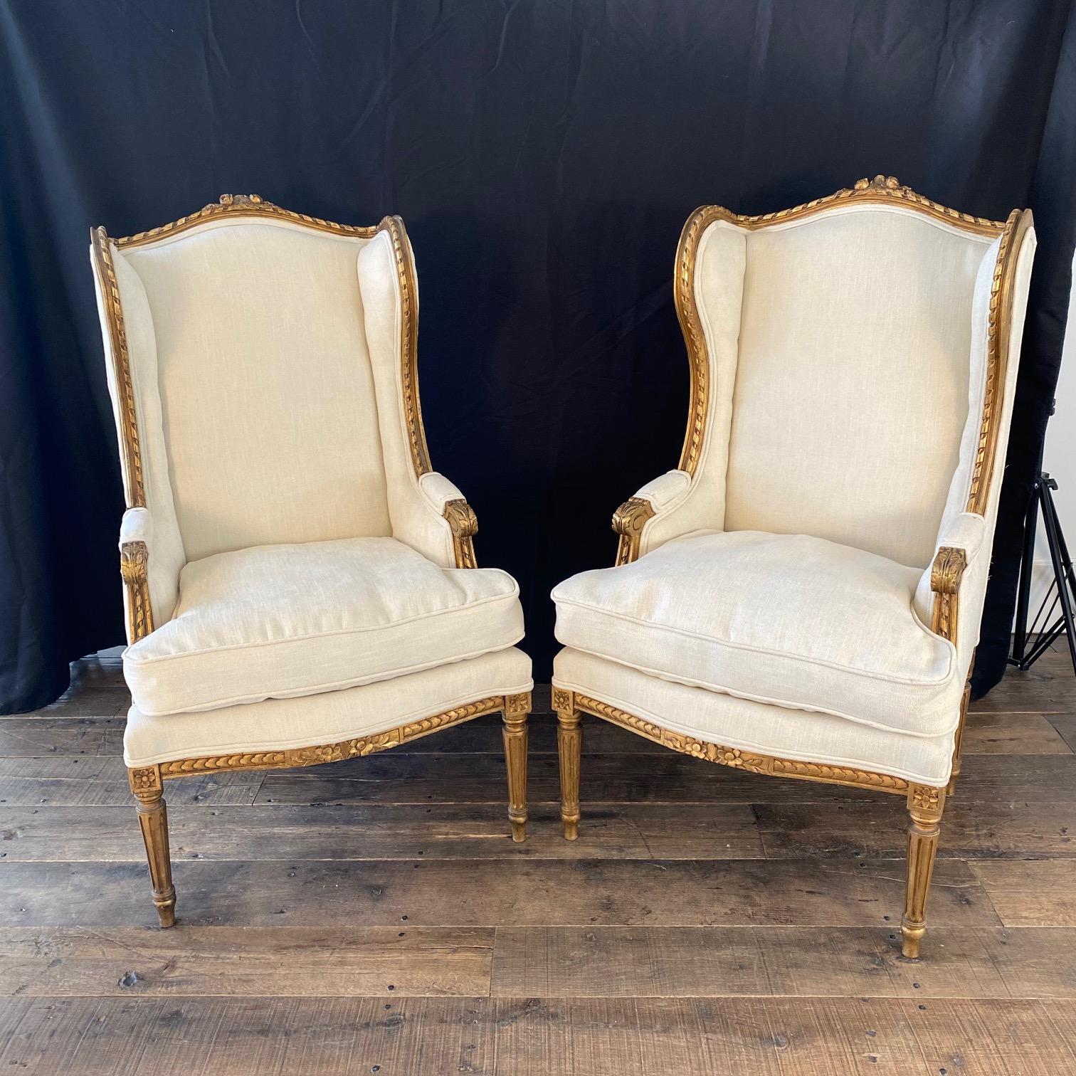 Elegant pair of French Louis XVI gold and off white giltwood wingback armchairs or bergere chairs from the 19th Century, both with high end new upholstery. The framework with arched seatback topped with flowers and foliates carved right into the