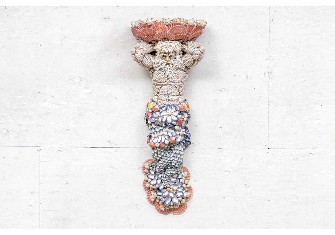 A powerful Triton or Neptune style figure in Caryatid form by Nantucket Shell artist Anne Jayson.  A tall elaborate shell encrusted wall decoration featuring a merman atlas figure with whiskers supporting the pink Scallop Shell Demi lune form top. A