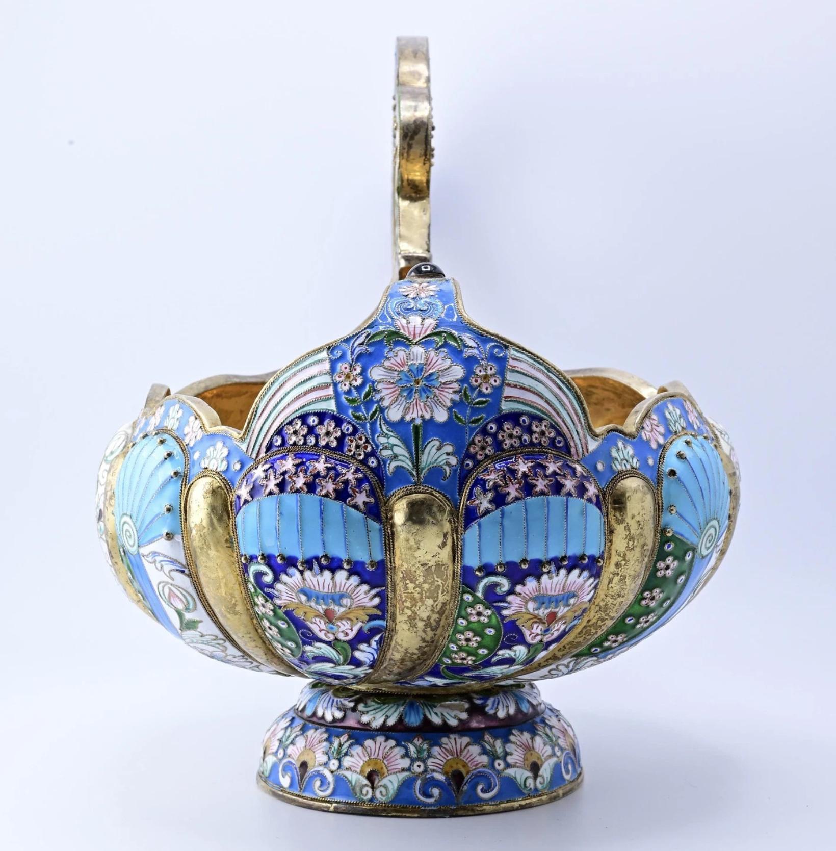 Esta vasija kovsh ornamentada rusa presenta un vibrante diseño con un intrincado trabajo de esmalte. El cuerpo está adornado con motivos florales y geométricos en tonos azules, verdes y blancos, acentuados con detalles dorados. El asa tiene una