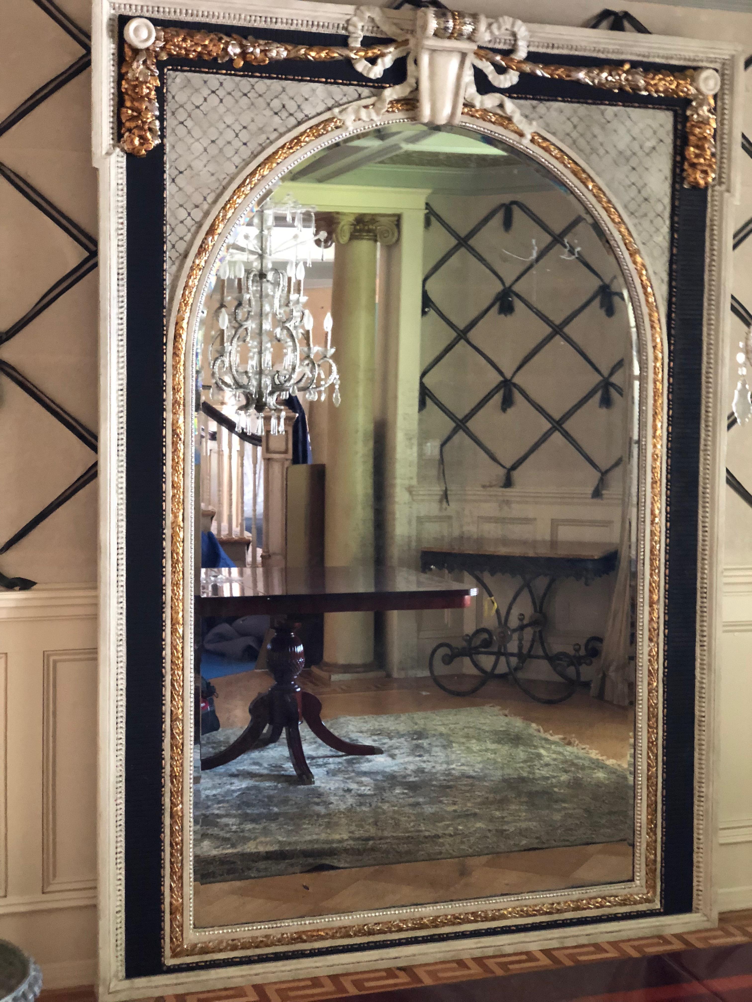 A luxe Louis XVI style mirror, monumental in size and destined to be the focal point of any interior. The grand arched beveled glass mirror is framed in wood, hand painted in 2012 with black, gold and white details. Original intricately carved