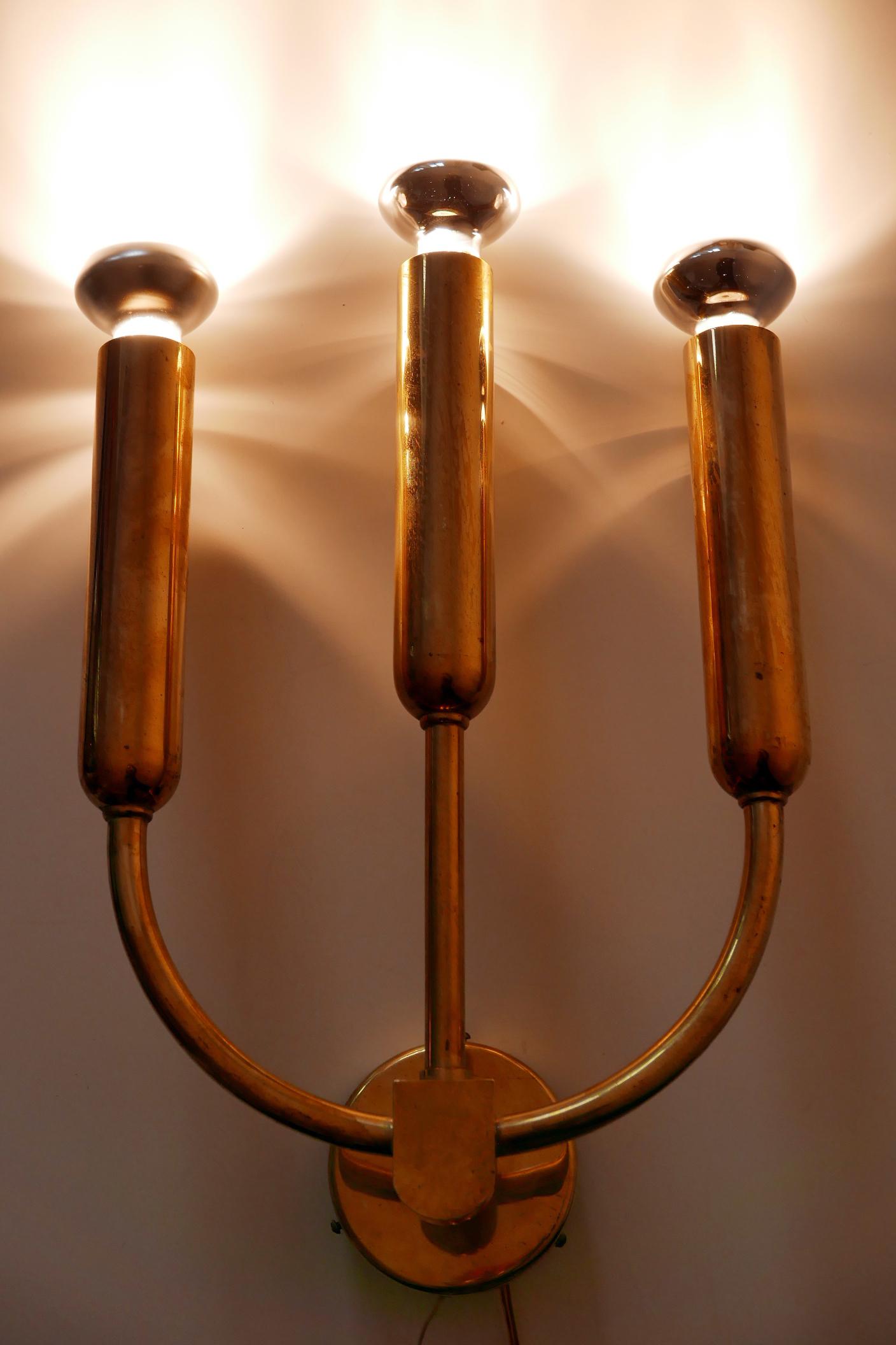 Exceptional, elegant and large Mid-Century Modern three-flamed brass wall lamp or sconce. Designed and manufactured probably in Germany, 1950s.

Executed in brass sheet and tubes, it needs 3 x E27 / E26 Edison screw fit bulbs. It is wired, in