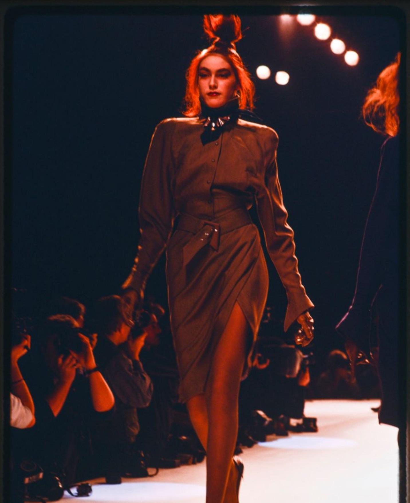 From Thierry Mugler Fall/Winter 1988 'Les Infernales' collection, variations of this black wrap-style dress debuted on the season's runway and features a sharp, structured silhouette. The design includes a high buttoned collar, long sleeves with