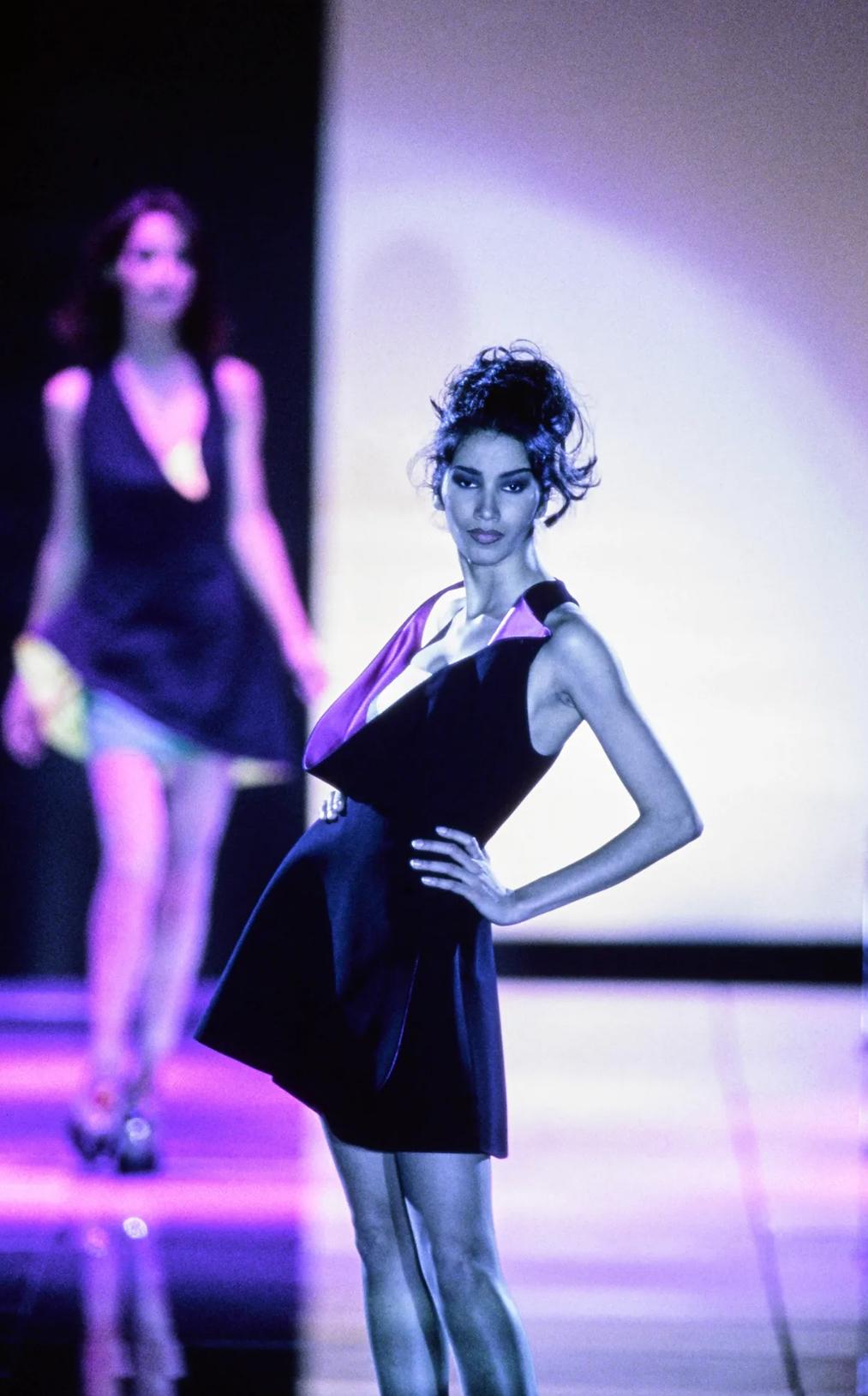 Cette fabuleuse mini robe enveloppante noire a été conçue par Gianni Versace. Issue de la collection Automne/Hiver 1991, cette robe a fait ses débuts sur le podium de la saison dans le cadre du look 80, porté par Gurmit Kaur. Cette petite robe noire