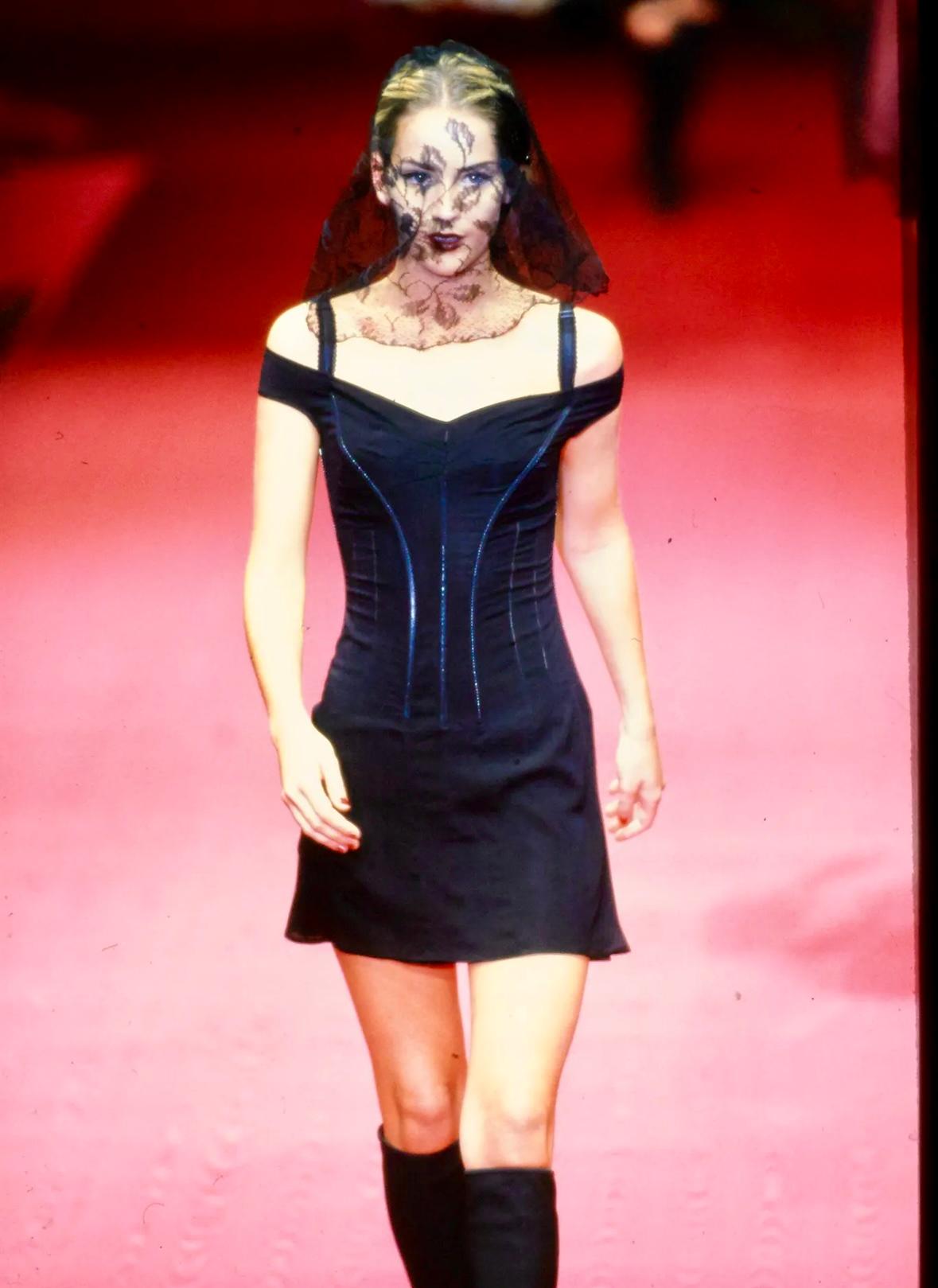Este vestido de noche Dolce & Gabbana Otoño/Invierno 1997 presenta un corpiño encorsetado con costuras contorneadas que realzan la silueta. Una superposición negra transparente se prolonga en una falda hasta el suelo, dejando ver un slip ajustado