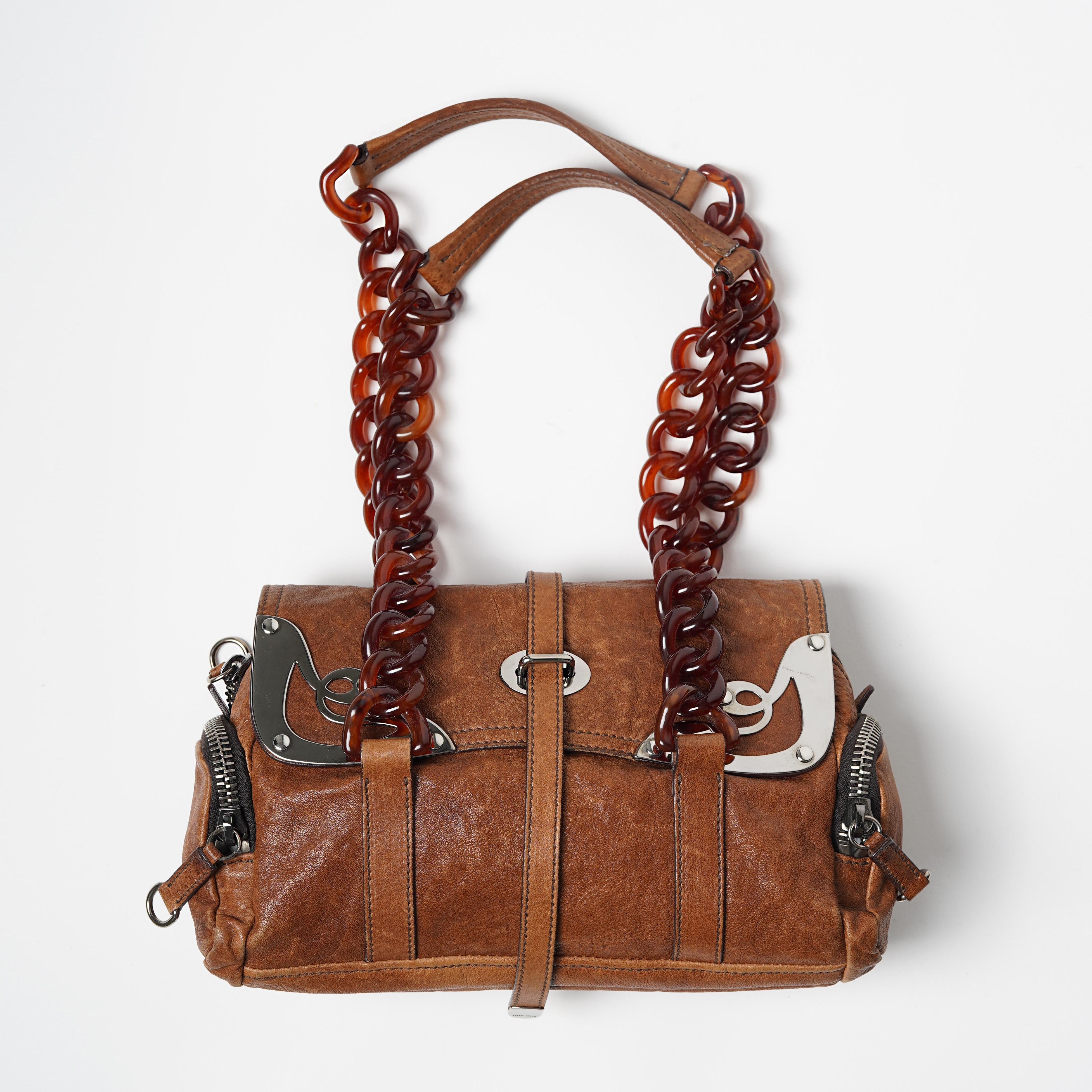 Ti presentiamo una borsa Miu Miu Runway Autunno/Inverno 2004 in pelle con ornamenti metallici distintivi, tasche con zip e manico a catena in celluloide. La borsa è stata vista sul look 7 della passerella di una sfilata in cui Miuccia Prada riteneva