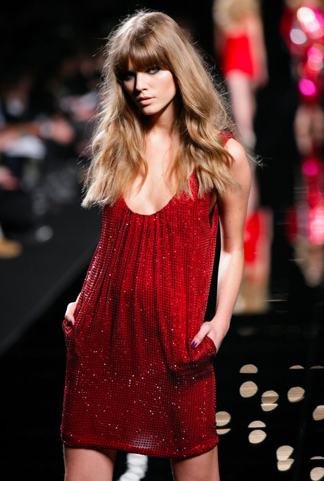 Este minivestido rojo rubí pertenece a la colección Otoño/Invierno 2007 de Emanuel Ungaro, diseñada por Peter Dundas. Presenta una silueta drapeada adornada con brillantes adornos de strass, un bolsillo en cada cadera y un profundo escote redondo.