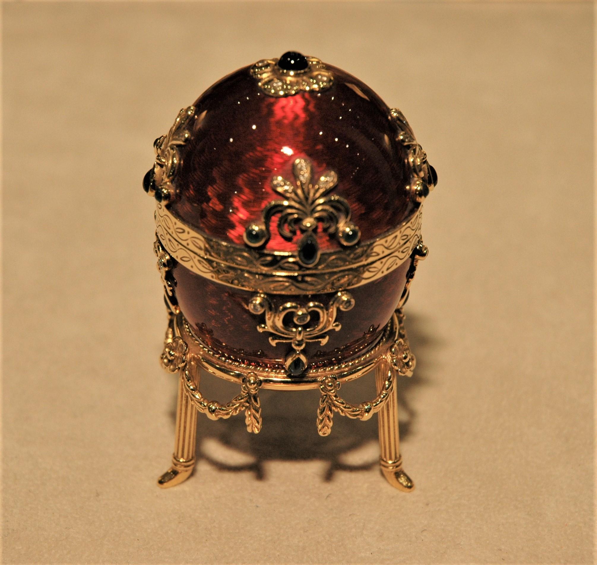 Seltenes Fabergé-Gold-Ei mit Goldständer. Es ist mit roter Guilloche-Emaille und Edelsteinen verziert: Smaragde (0,40 Karat), Saphire (3,15 Karat), Diamanten (0,56 Karat). Sie ist zu öffnen und man kann einen speziellen Ring hineinstecken. Es ist