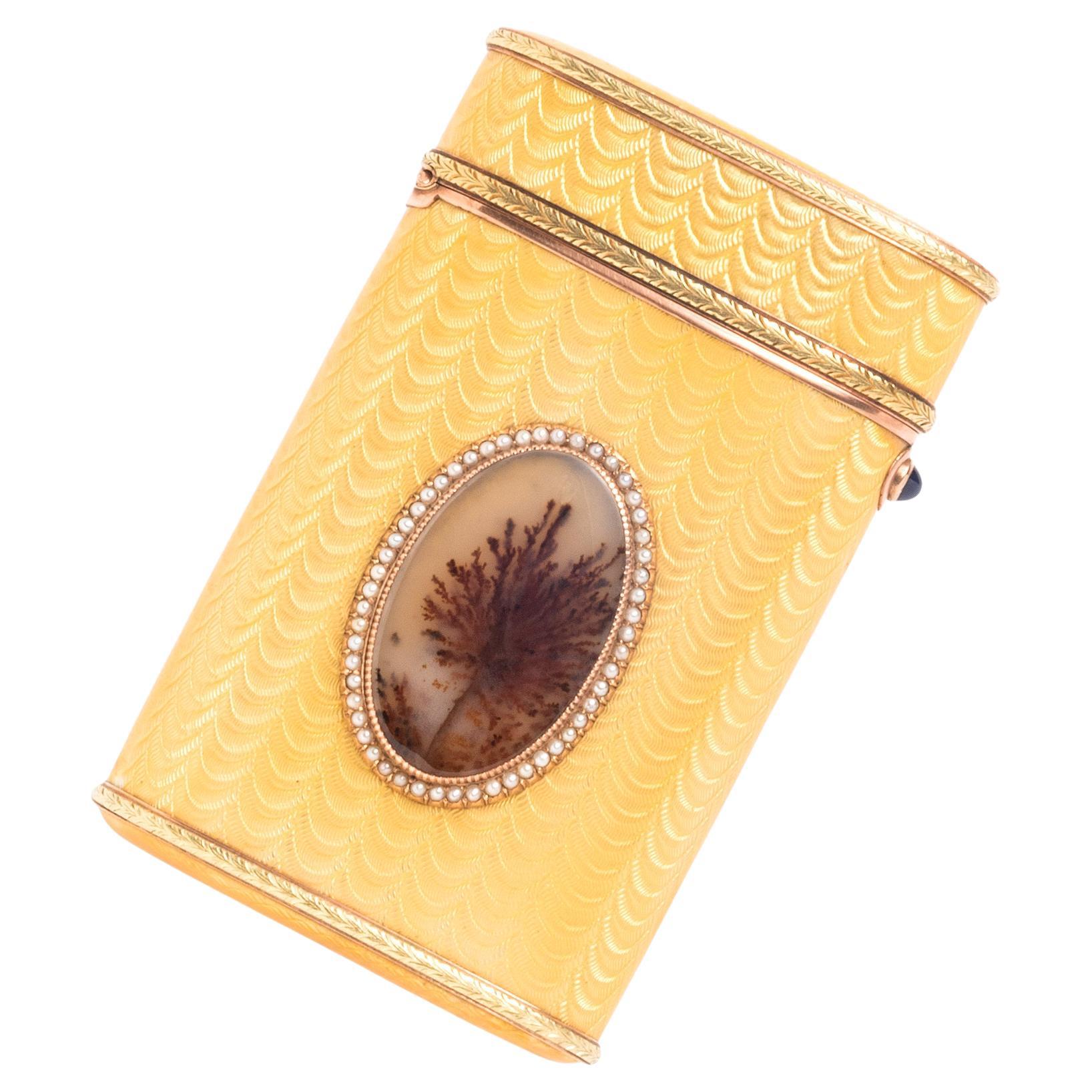 This bicolor gold and enamel cigarette case by Fabergé features an applied moss agate oval decoration set within a seed pearl and gold border. The body is decorated with yellow guilloche enamel and applied laurel bands at the base and cover. The