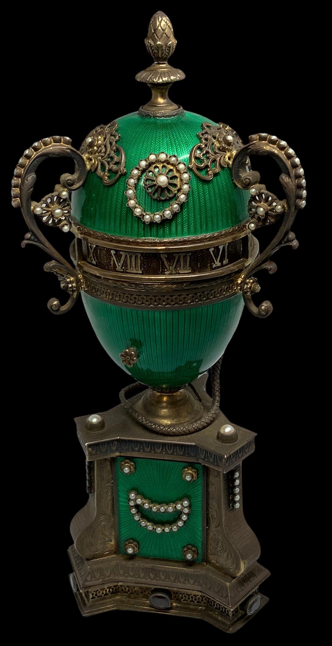 An emerald color guilloche egg adorned with decorative silver flowers and three scrolls handles of acanthus leaves encrusted with cultured pearls. A serpent is coil around the bottom base of the egg and his garnet head/tongue pointing to the hour.