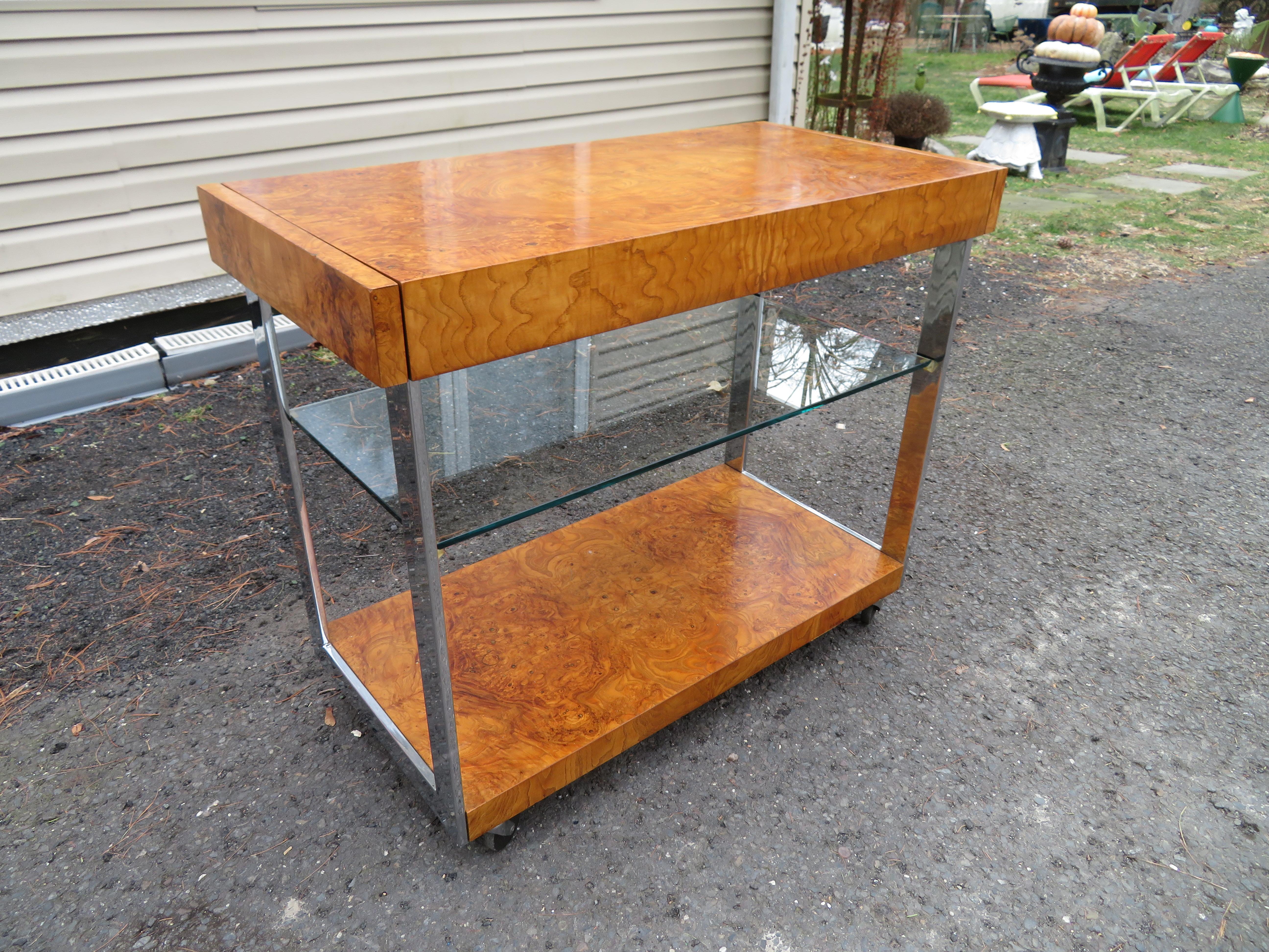This is a beautiful bar cart in chrome and burl wood in the style of Milo Baughman. It is part of the Alpha II by Lane circa 1975. Veneer burl wood shelves are connected to polished chrome flat bars. The cart or trolley is supported by castors and