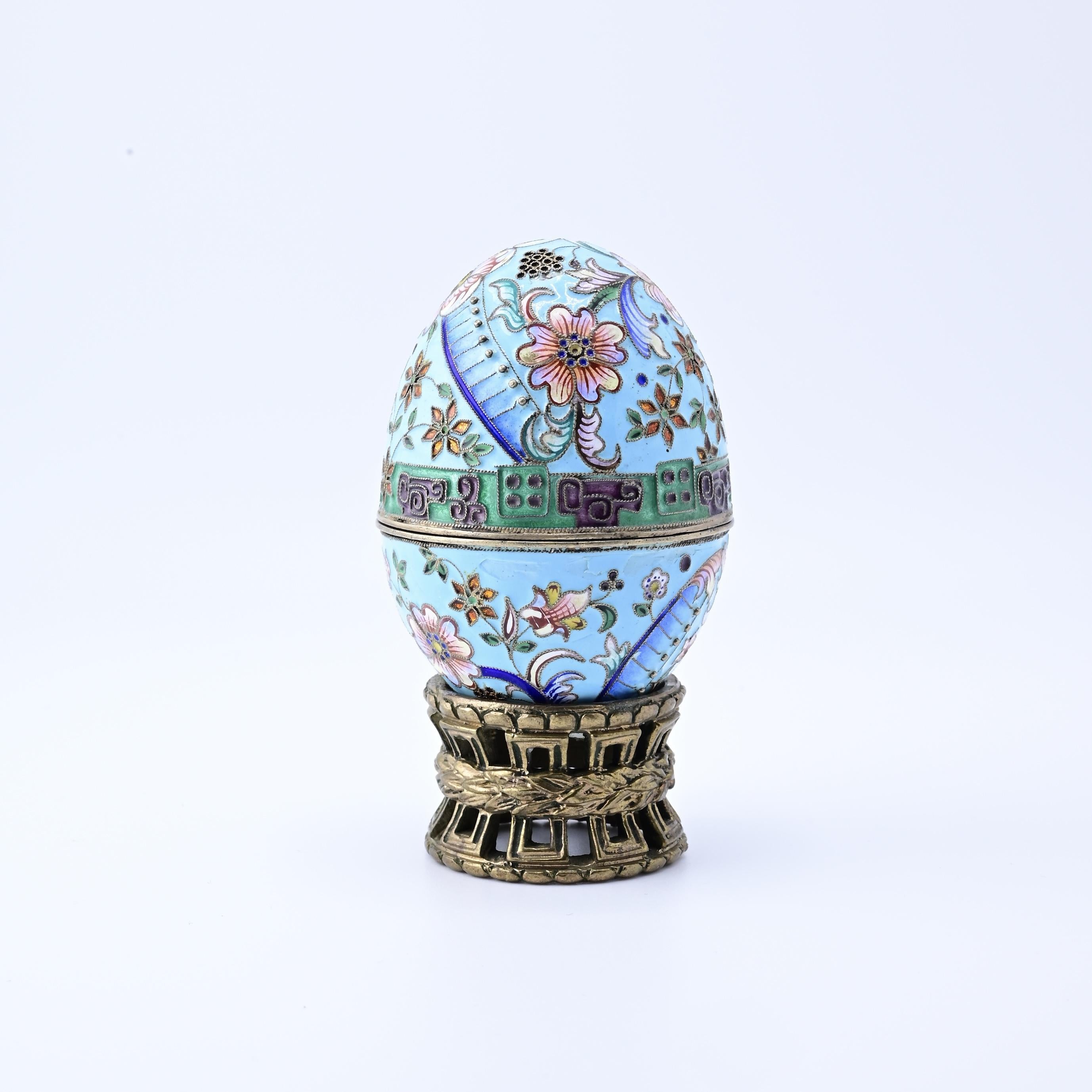 Este fabuloso huevo decorativo ruso de plata esmaltada es una cautivadora obra de arte. Adornado con intrincados motivos florales, el huevo presenta un delicado fondo azul con vibrantes flores y hojas. El diseño se acentúa con una banda de motivos