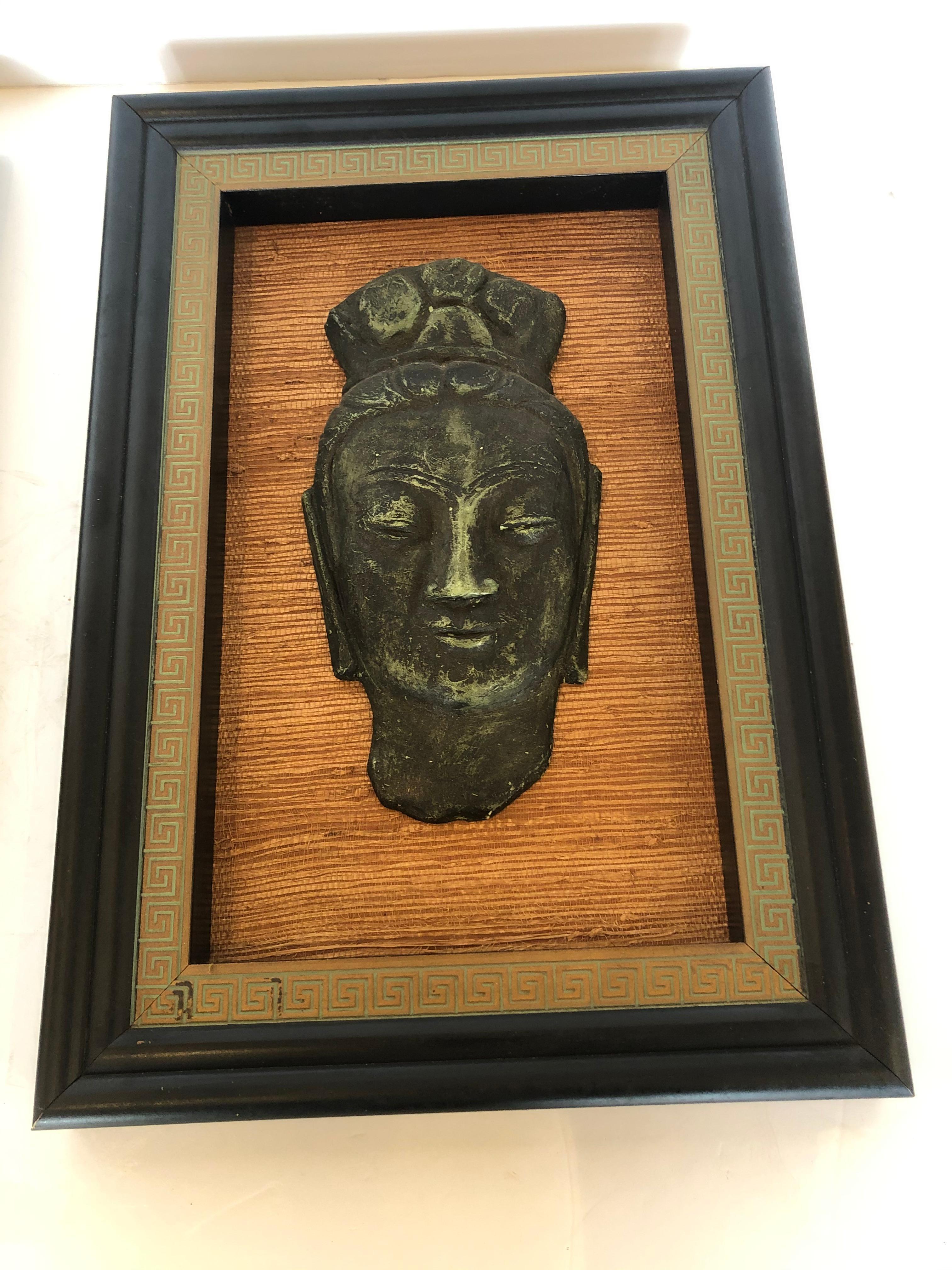 A superb pair of Hollywood Regency sculptural relief Buddha heads mounted on grasscloth backgrounds and framed stylishly in ebonized frames with gold and celadon green Greek key borders. Tony Duquette would have loved these.