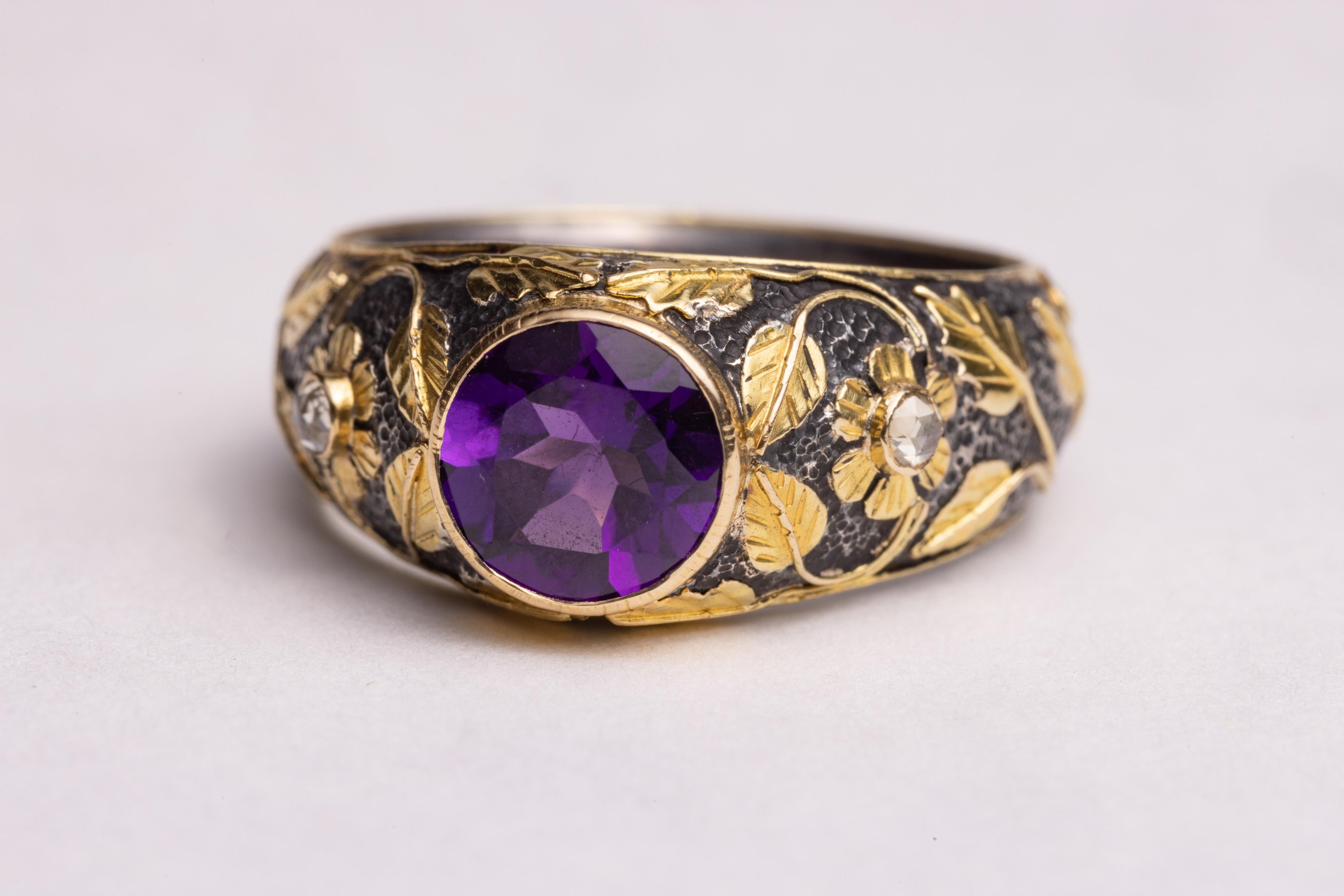 An unusual ring in a combination of oxidized sterling silver overlaid with 18K gold in a floral vine pattern all hand-tooled.  It features a stunning, round faceted amethyst in the center with two round, faceted diamonds on the side in the center of