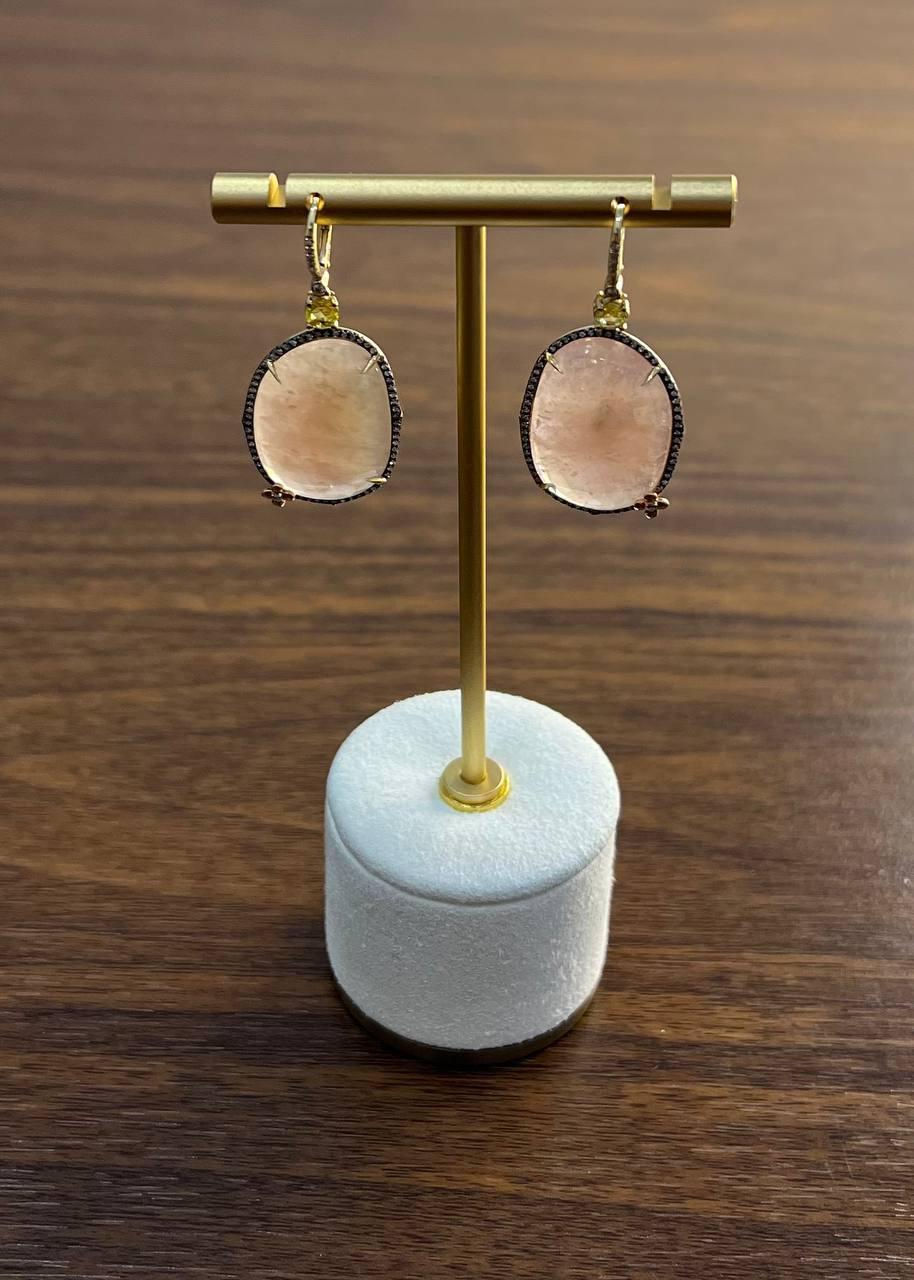 A pair of elegant gemstone earrings. Each earring features a large, faceted oval pink peach quartz with a soft pink hue. Bordered with a delicate line of small, sparkling diamonds, in a 14K yellow gold frame. The lever-back clasps are adorned with a