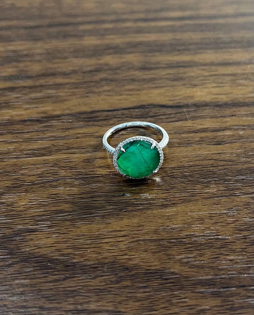 Ring with a large green emerald in faceted cabochon set in 14k white gold. The stone is faceted and surrounded by a halo of small clear diamonds. The band features intricate detailing.