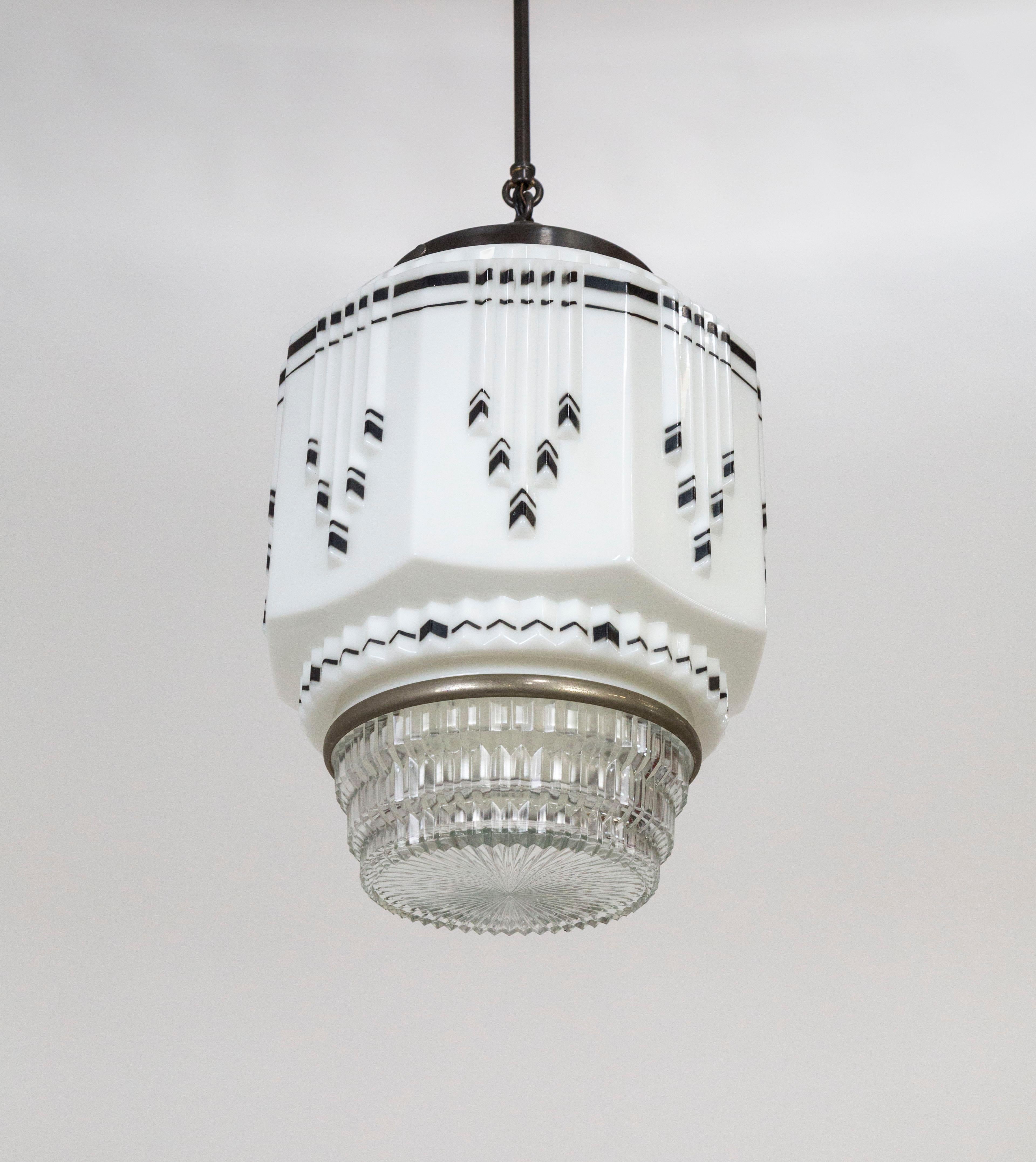 An Art Deco, cylindrical pendant light made of faceted, ridged, white, molded glass, accented with black stenciled zig-zags; with a chrome-rimmed base of clear, stepped, pressed glass with a radial texture. Held with a long, slender, brass stem and