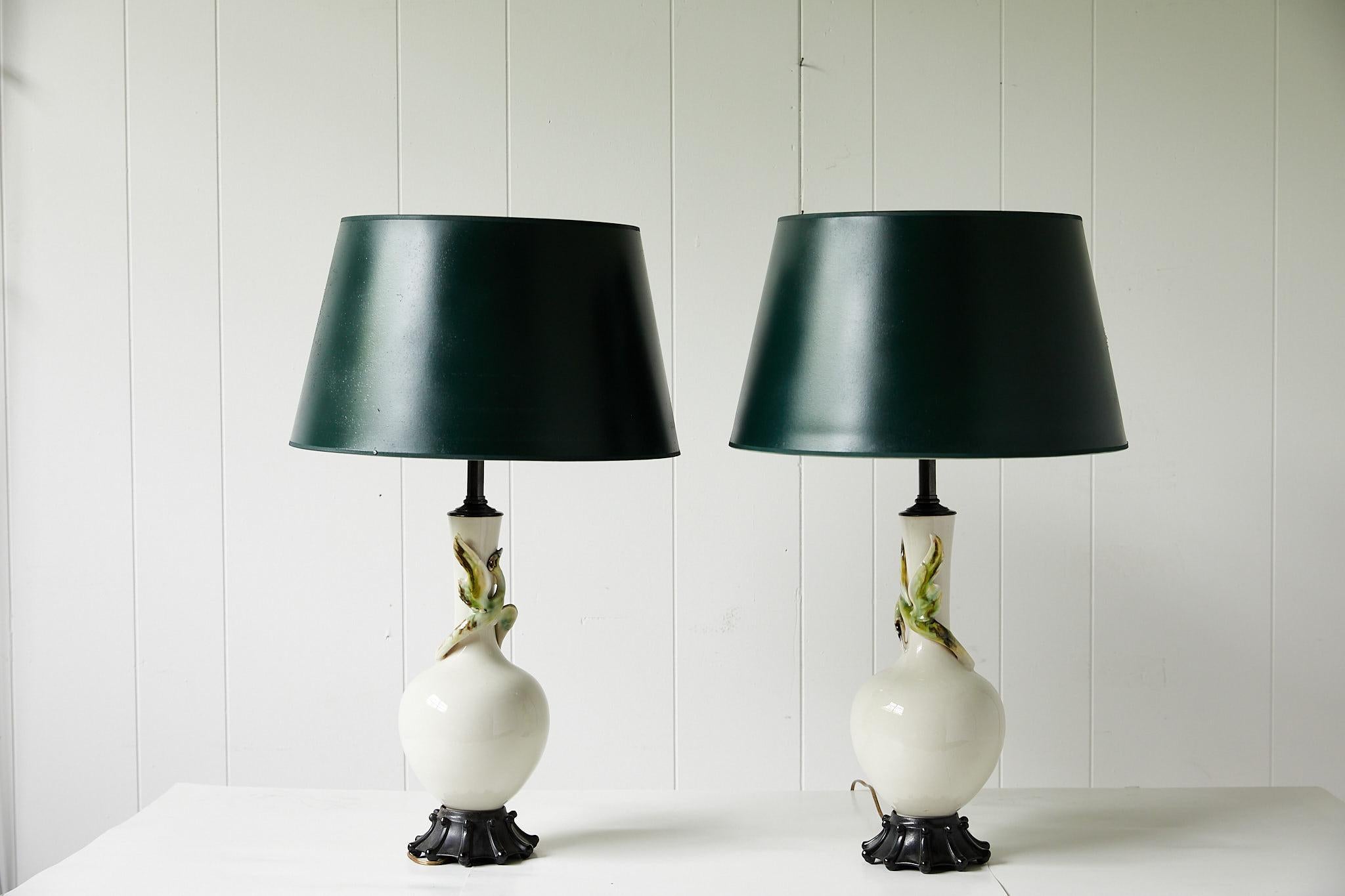Early 20th century facing pair of lamps made of cream ceramic and in a simple vase form. Each lamp has a slightly different ceramic bird of green and brown tones decorating it's neck. The lamps are mounted on carved wooden bases and topped with