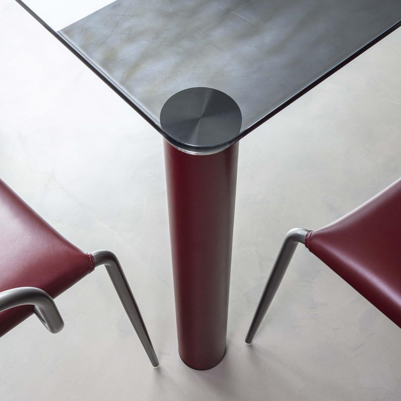 Zylindrische Stahlbeine, luxuriös mit burgunderrotem Leder überzogen, bilden den Sockel dieses majestätischen Tisches, eine hervorragende Ergänzung für moderne Esszimmer. Eine kostbare Tischplatte aus Rauchkristall mit abgerundeten Ecken und