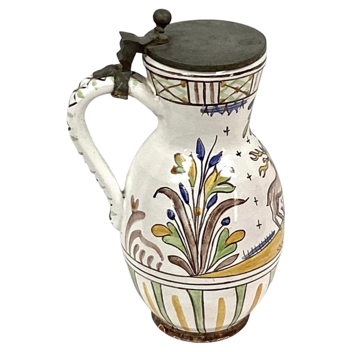 German Faience pitcher/jug with hinged pewter lid and handle. Hand painted floral design, along with stag and dog, on white background. Top of lid is monogrammed. Has minor wear from lid on rim of jar and on bottom. Inside is in excellent condition