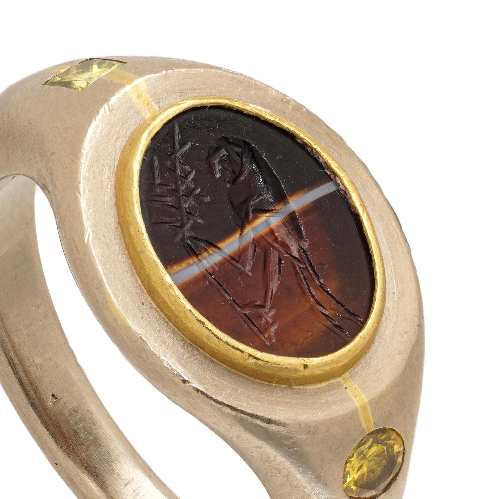 A distinctive 18kt white and yellow gold ring by German goldsmith Falco Marx, featuring a carved Roman intaglio and two yellow diamonds. The oval layered agate intaglio (11 × 8 mm), bezel-set in yellow gold, depicts a stylised bird perched beneath