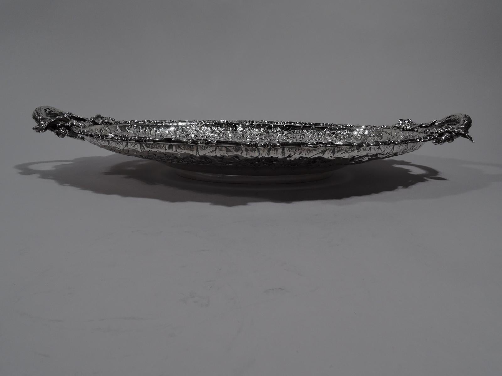 Fancy repousse sterling silver serving platter. Made by Tiffany & Co. in New York. Plain oval well with leaf-and-dart border. Wide tapering shoulder with flowers and ferns on stippled ground. Rim has applied scrolls and flowers. C-scroll end branch