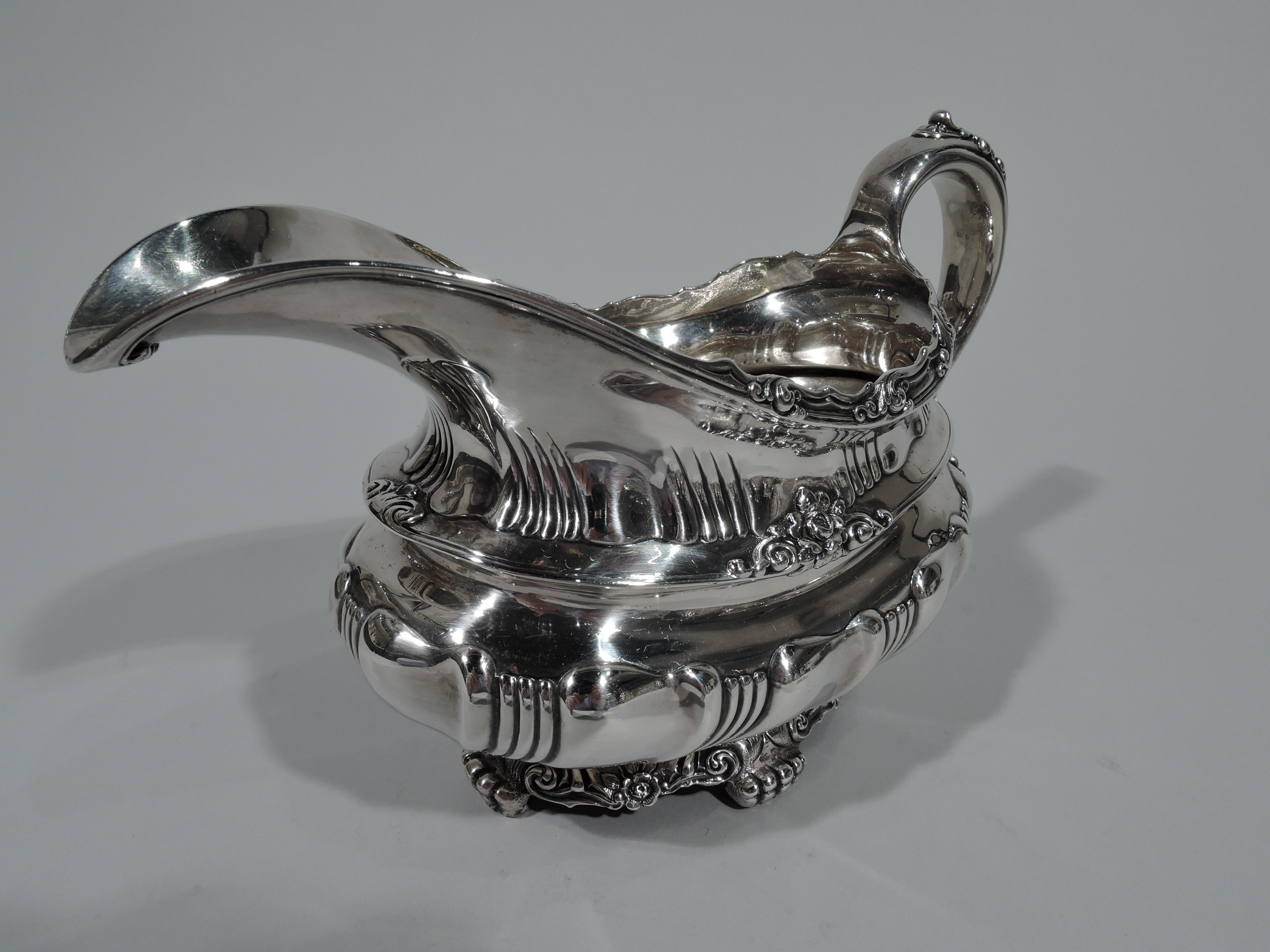 Fancy sterling silver gravy boat. Made by Tiffany & Co. in New York. Bellied oval with helmet mouth, leaf-capped high-looping scroll handle, and foot ring with 4 paw supports. Alternating scrolled fluting and ribbing. Chased flowers and leafy