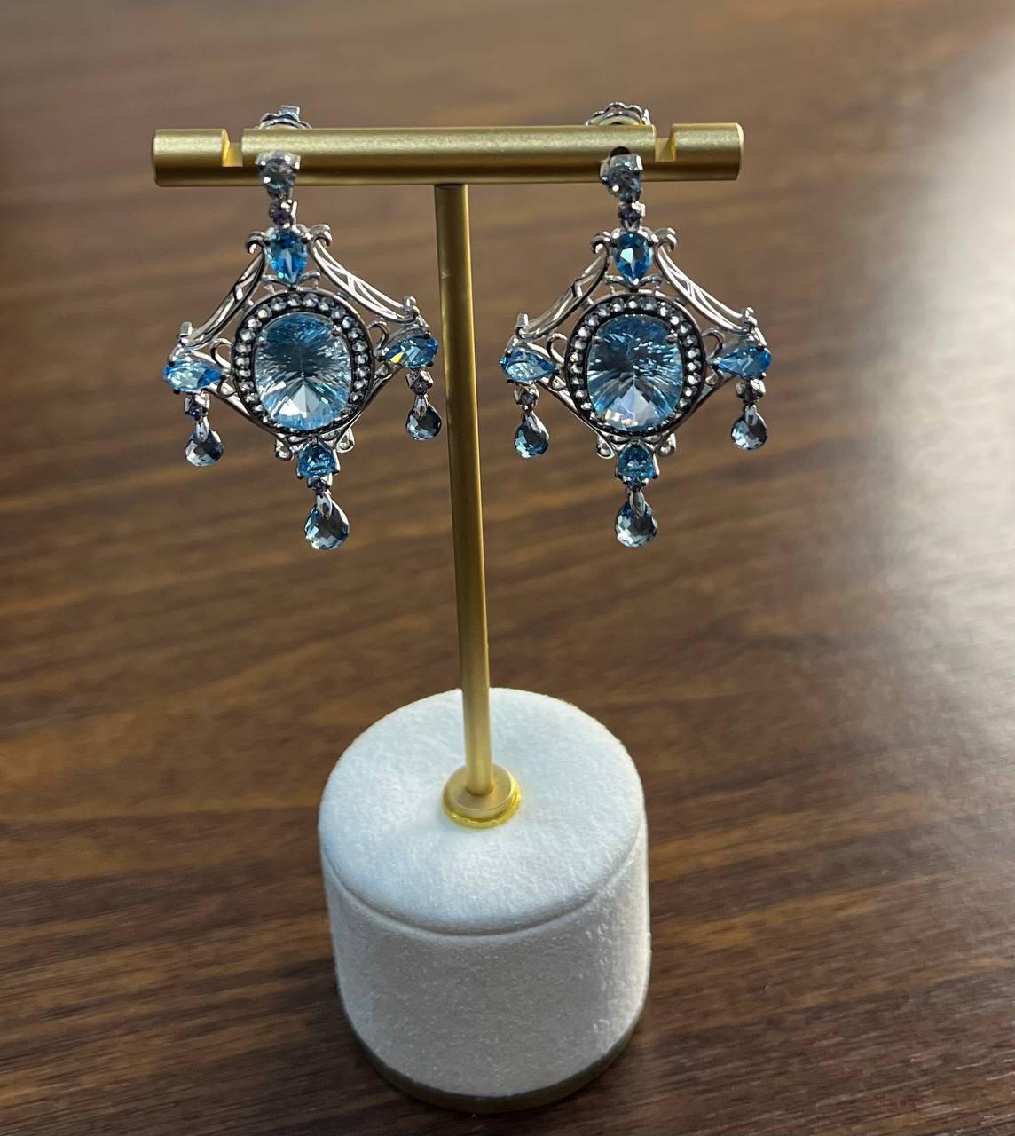 Exquisite dangle earrings featuring intricate designs with shimmering blue topazes. The central stones are encircled by delicate 14K White gold metalwork, adorned with smaller teardrop-shaped topazes and diamonds that cascade gracefully.

Elegant