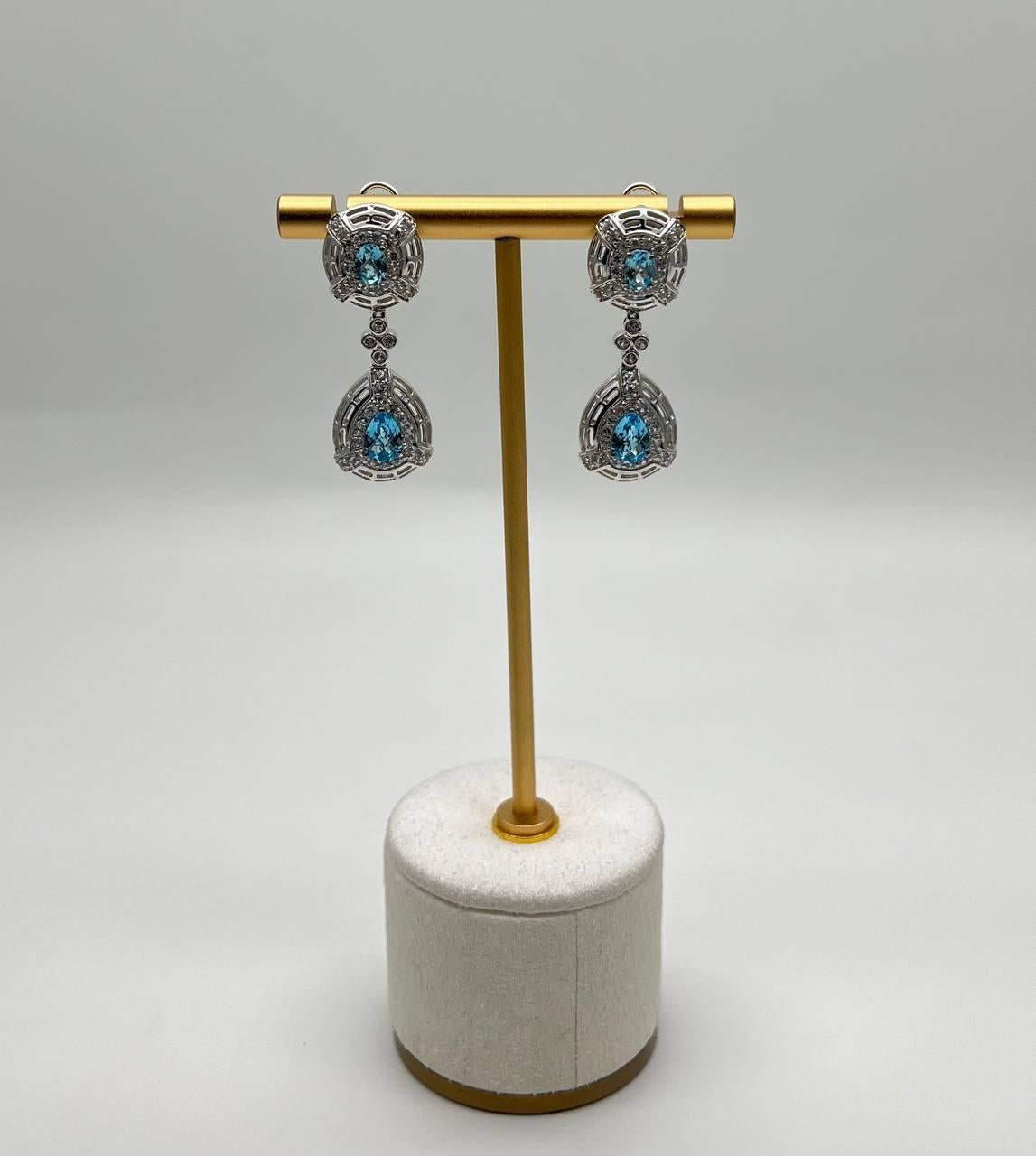 These drop earrings feature intricate 14k white gold metalwork with vibrant blue topazes. The design includes a teardrop shape, accented by detailed lattice patterns. The earrings are suspended from a simple omega lock, allowing for elegant