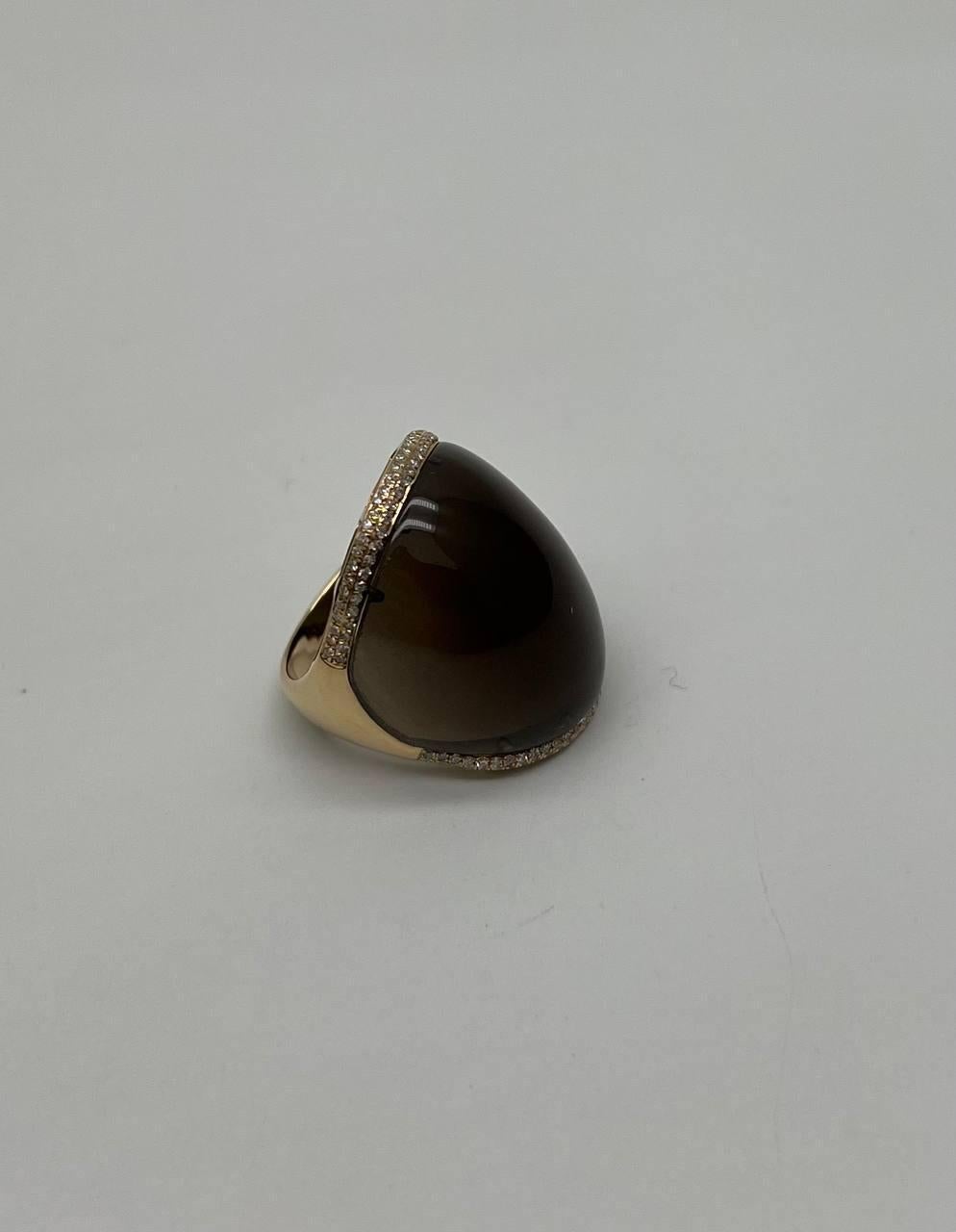Questo pezzo impressionante presenta un quarzo fumé cabochon, liscio e audace, con una ricca e profonda tonalità marrone. La pietra è incastonata in un'ampia fascia, accentuata da un delicato bordo di diamanti scintillanti che aggiungono un tocco di