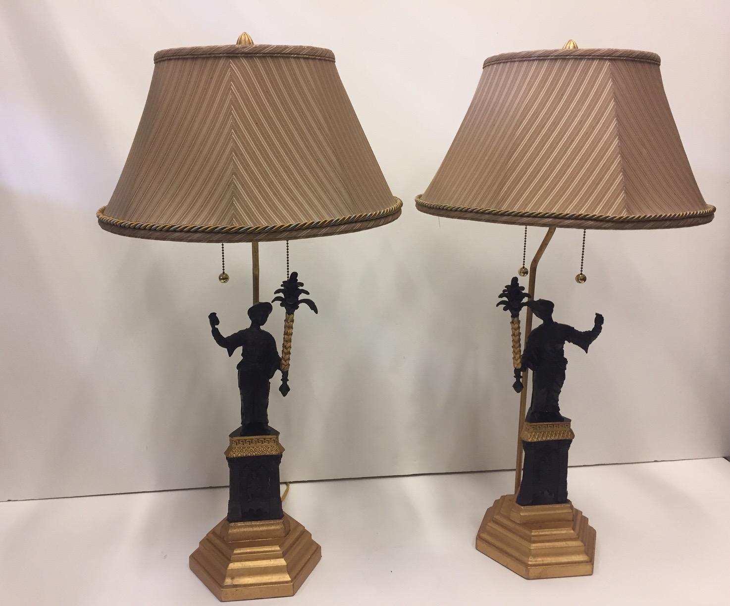 A gorgeous pair of designer patinated bronze figural lamps on giltwood bases having period dressed figures holding stylized torch like objects. Beautiful gilded details on the torch and bases under their feet, including Greek key embellishments.