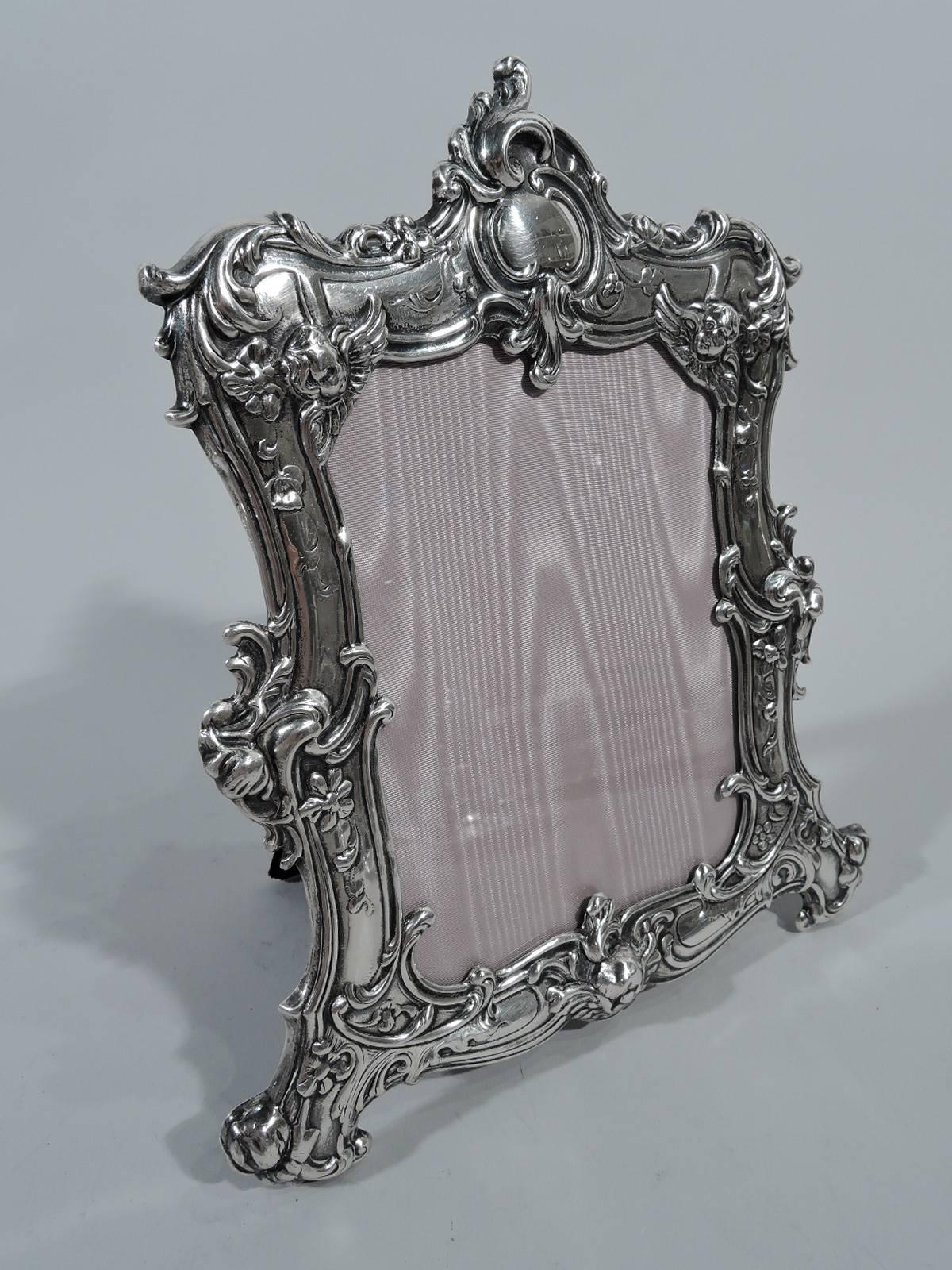 Fancy Rococo sterling silver picture frame. Made by Gorham in Providence. Dynamic and scrolled surround with bracket feet: Ornament chased and applied: Flowers, leaves, scrolls, and winged cherub’s heads. At top asymmetrical cartouche (vacant). With