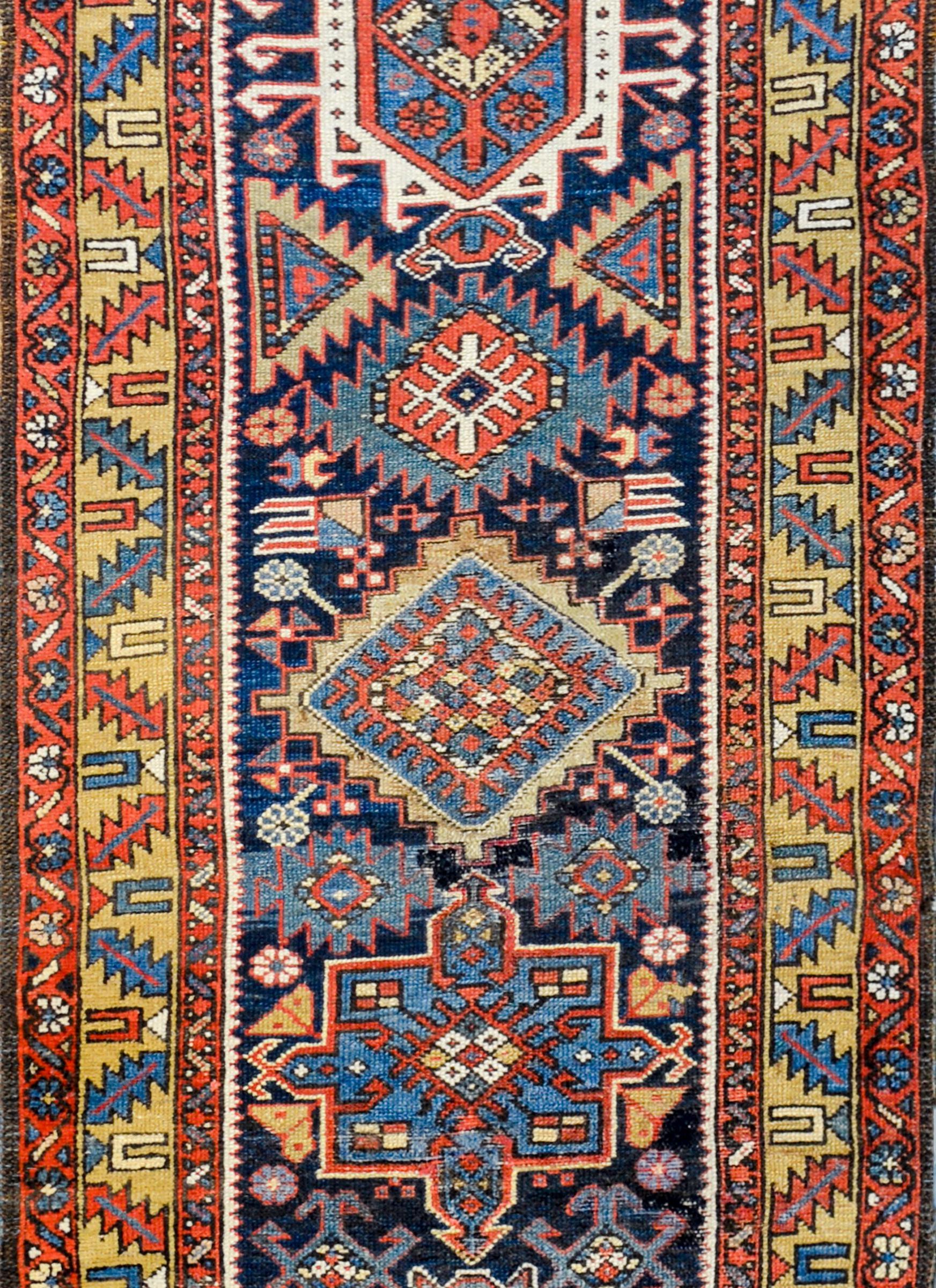 A fantastic early 20th century Persian Heriz runner with a bold tribal pattern consisting of multiple stylized floral medallions woven in gold, crimson, light indigo, and white, amidst a rich dark indigo field of more stylized flowers. The border is