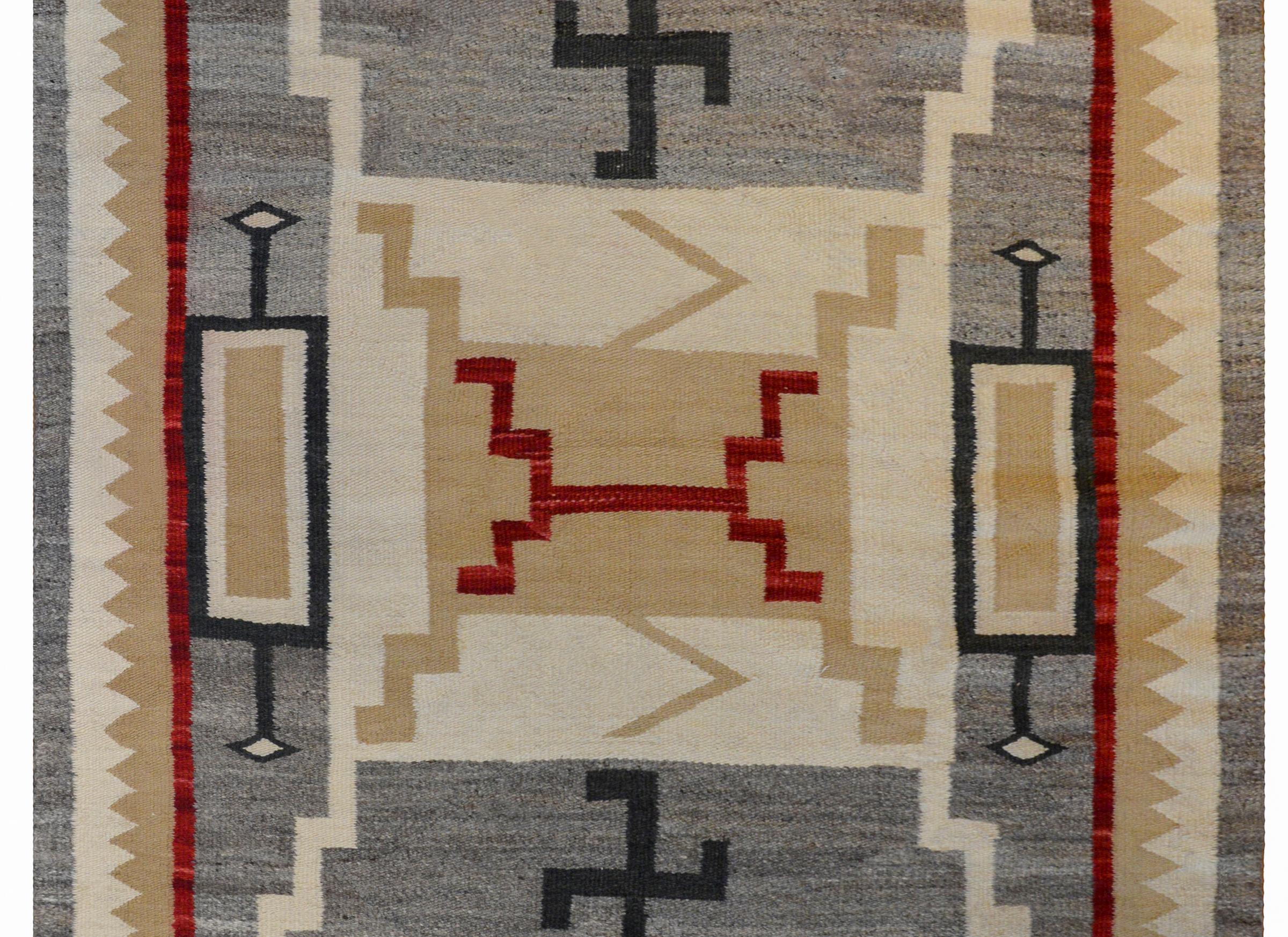 A fantastic early 20th century Navajo rug with a busy pattern containing multiple wide zigzag stripes woven in crimson, tan, and cream colored wool flanked by black swastikas, symbols of good luck, on a gray background surrounded by a wide tan and