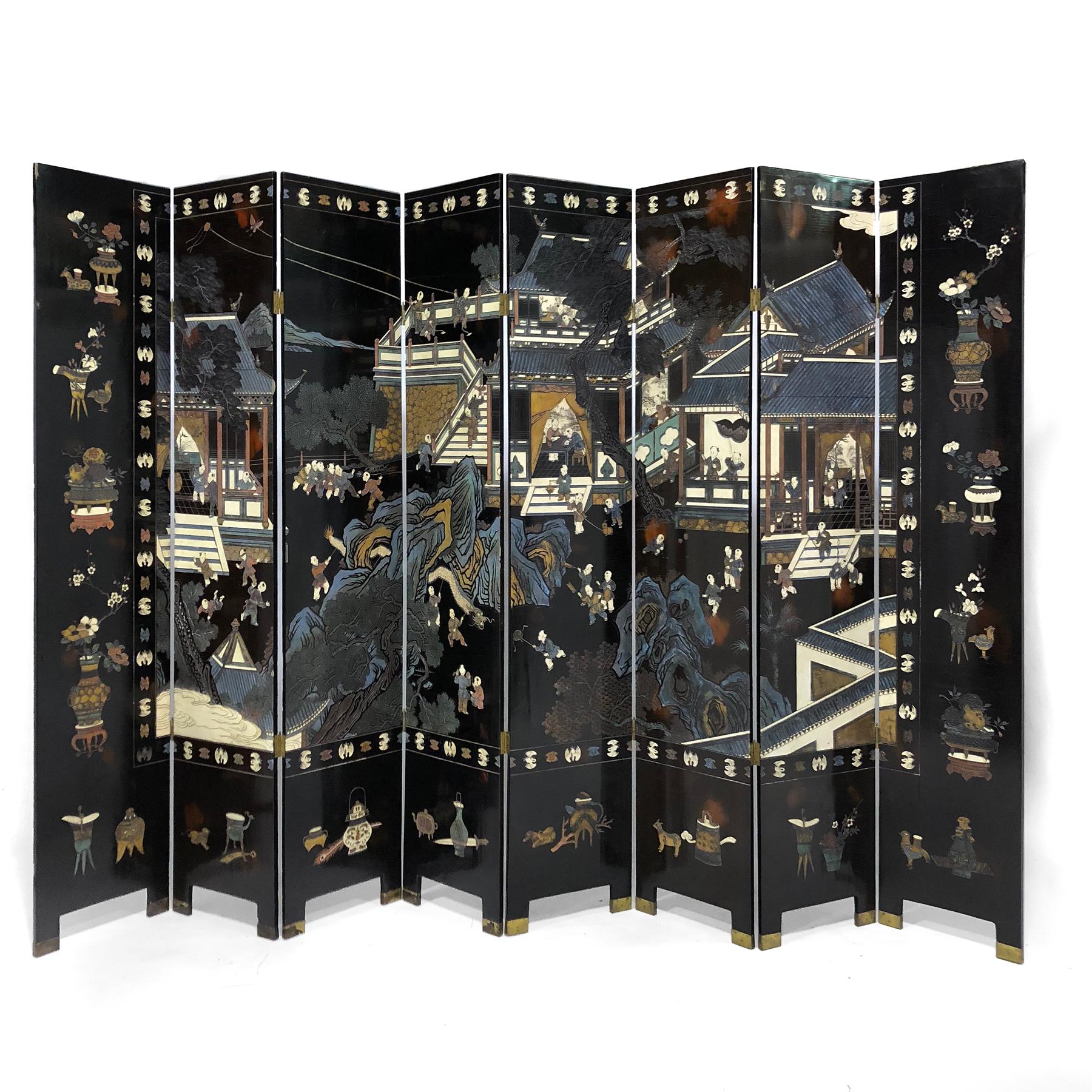 This screen has eight panels with brass feet. The front features a classic highly detailed village scene with figures in pavilions bordered by a pattern of cranes and flowers and scholar's objects or antiques framing the scene on three sides, while