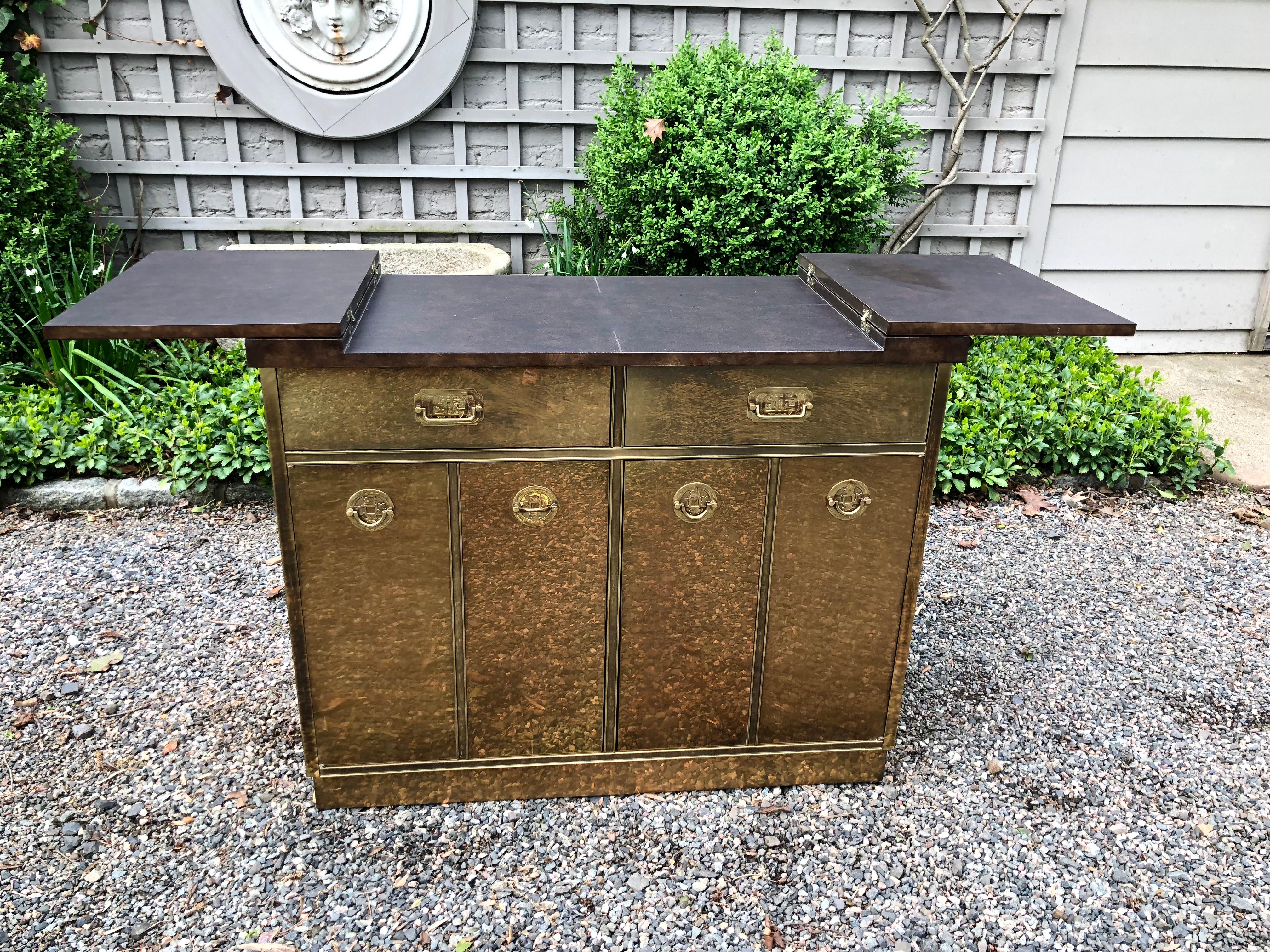 A rare midcentury brass clad bar and cabinet by Mastercraft. The bar features a lacquered faux wooden top that opens to a laminate surface suitable for serving drinks. The interior has two drawers and four shelves for storage. The bar has the iconic