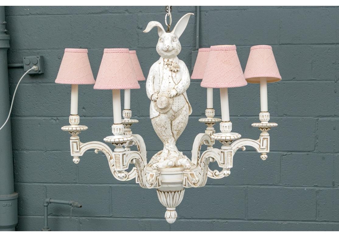 An intentionally distressed and worn finish in a carved wood six light chandelier in white paint with the figure of a dapper male Rabbit  as the support. He wears a weskit and jacket with a flower in the lapel and stands holding his hat cross-legged