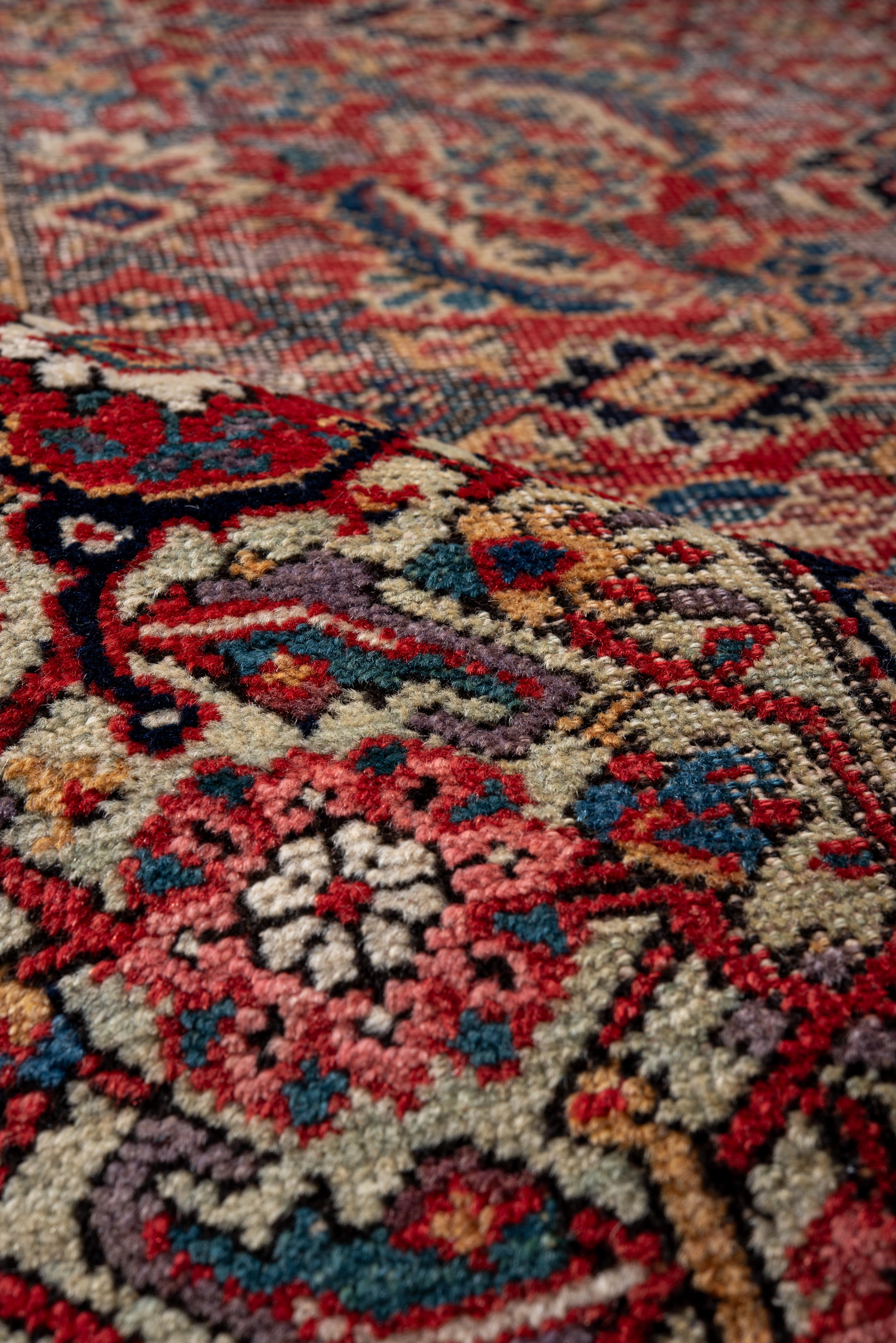 Dieser antike Farahan-Teppich besticht durch ein aufwändiges Allover-Muster aus Paisley-Boteh-Motiven, die mit Rosetten und zartem floralem Maßwerk verwoben sind, auf einem tief karminroten Feld. Elfenbeinfarbene und nachtblaue Bordüren sorgen für