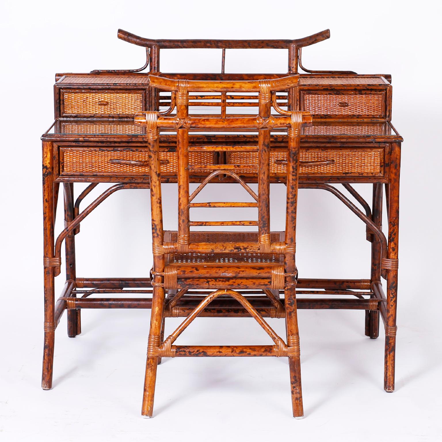 British colonial desk and caned chair with faux bamboo frame highlighted by a pagoda motif, two glove boxes, glass covered writing surface, two drawers, grasscloth panels, and splayed feet. Perfect fit for those looking for the hottest in the wicker