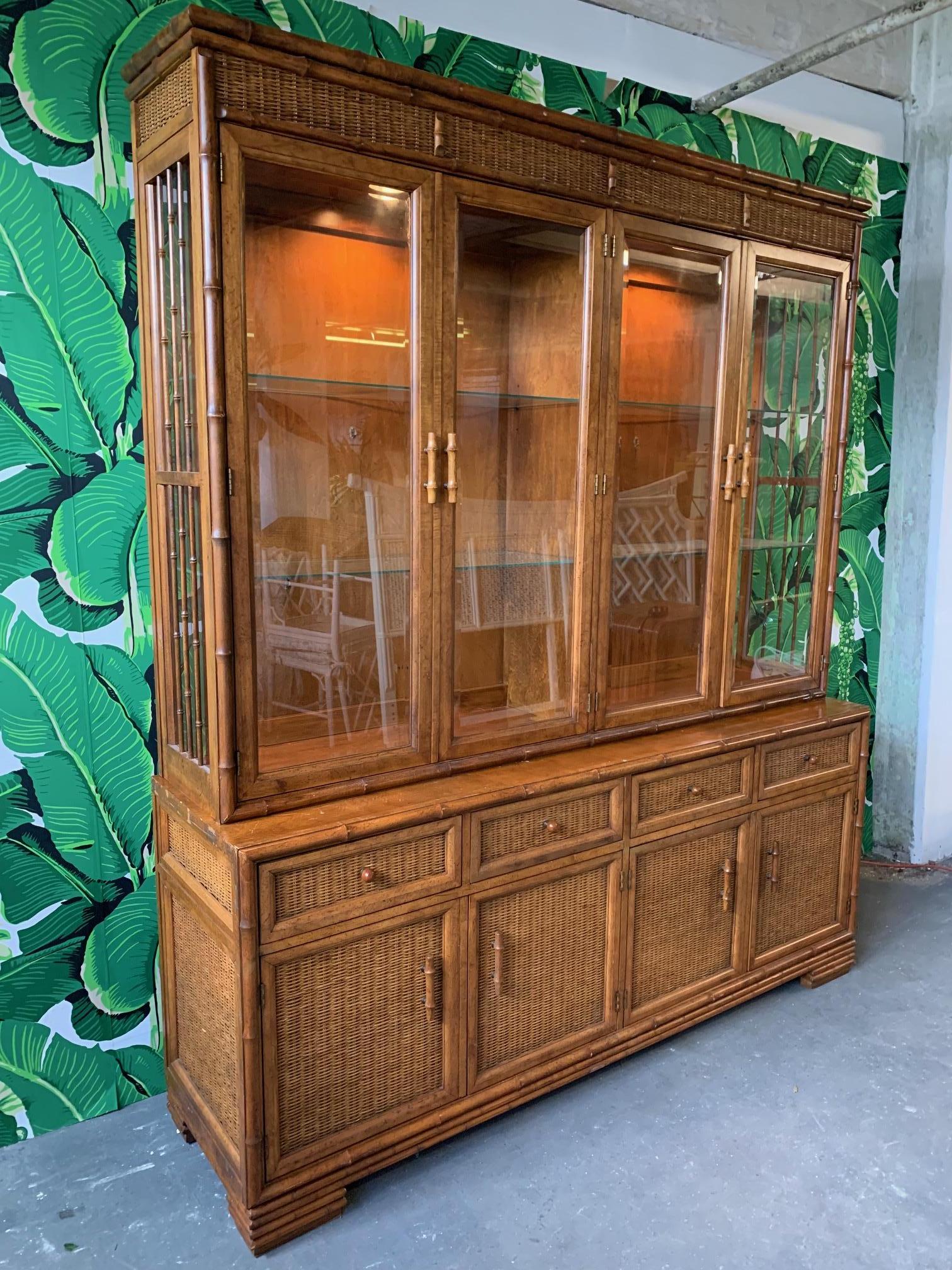 Large faux bamboo china cabinet features lighted display cabinet, glass shelving, and wicker detailing on door and drawer faces. Very good vintage condition with some abrasions on side (see photo). Cabinet is two pieces, top and bottom.
